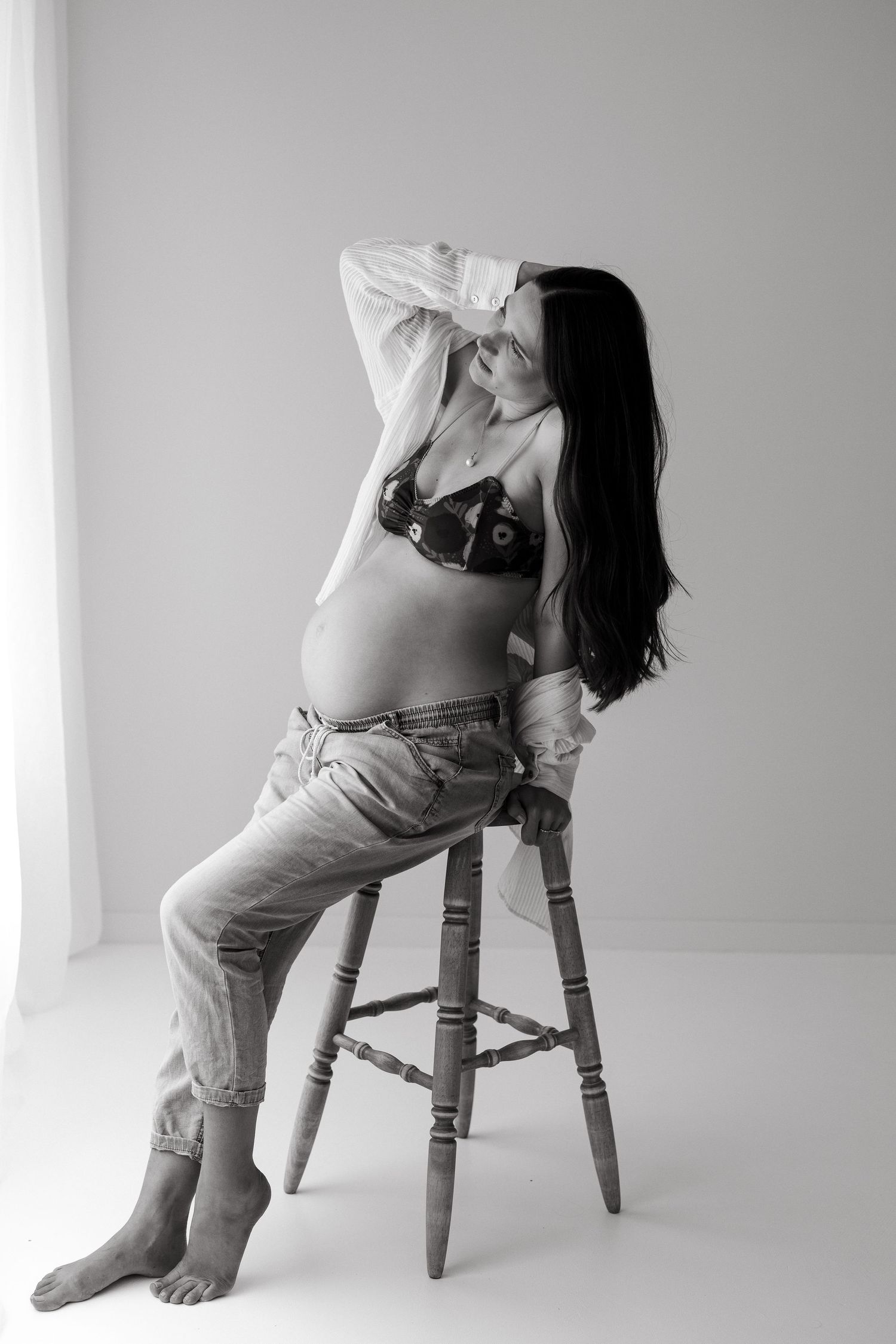 Maternity boudoir photo of pregnant woman posing on wooden stool in casual attire.
