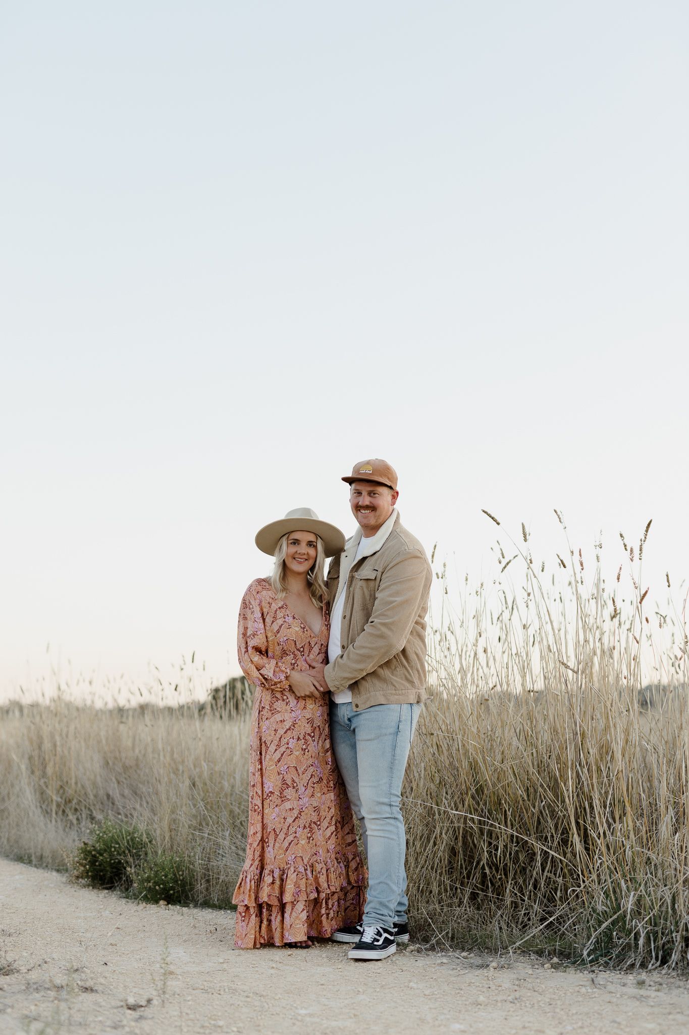 A couple in casual attire shares a romantic moment on a dirt path surrounded by tall grass at sunset.