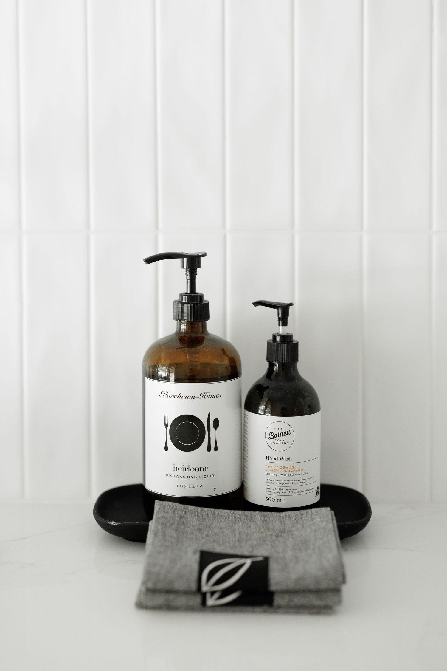 Minimalist bathroom set with amber glass pump bottles and gray folded towel on white surface against white paneled wall.