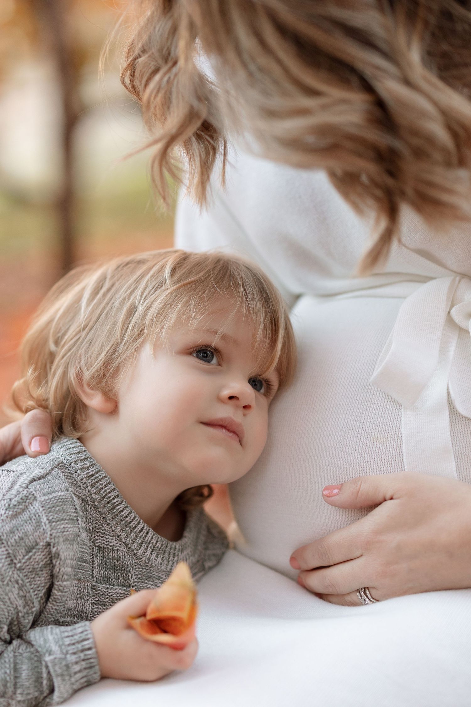 A tender motherhood moment with a toddler in a grey sweater resting against a white maternity dress.