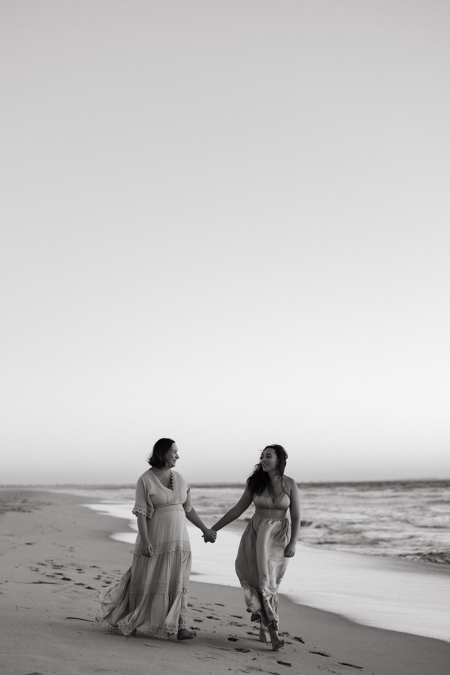 Two people walk hand in hand along a misty beach shoreline in black and white.