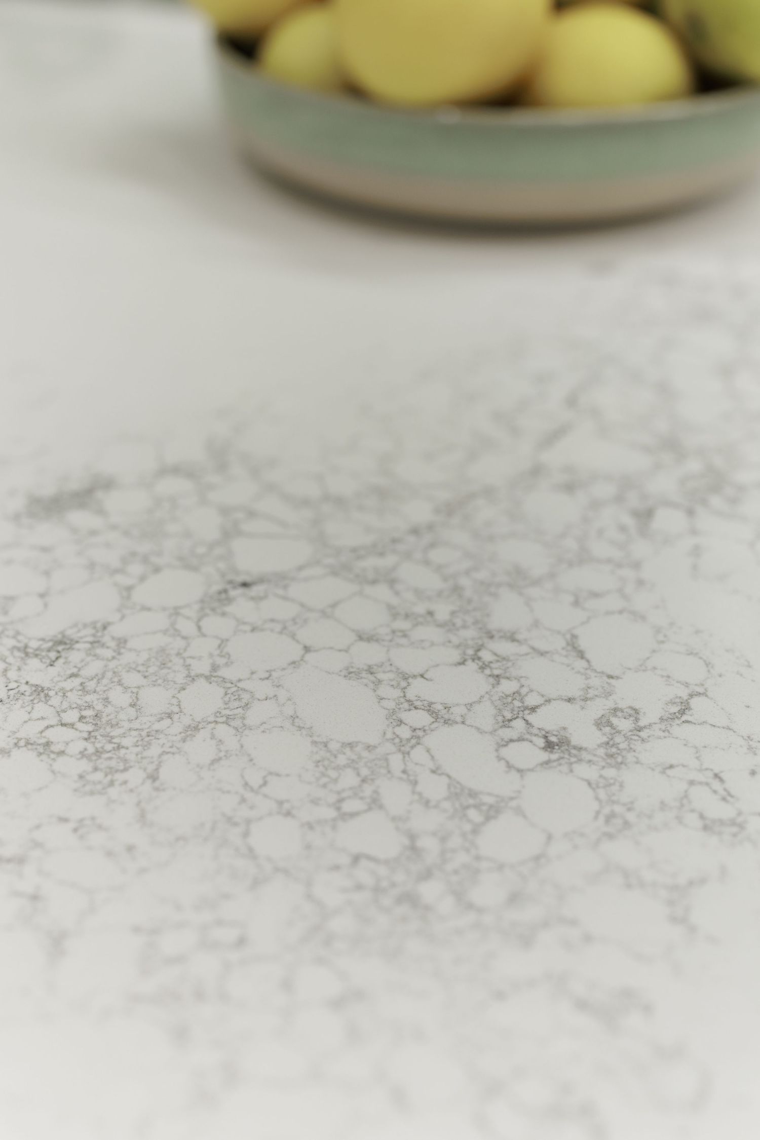 Close-up of a white marble countertop with grey veining, with a bowl of fresh lemons blurred in the background.