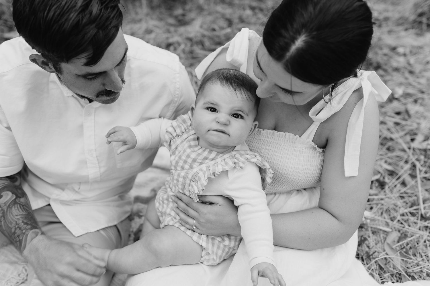 Black and white photo of parents embracing baby wearing checkered outfit outdoors.