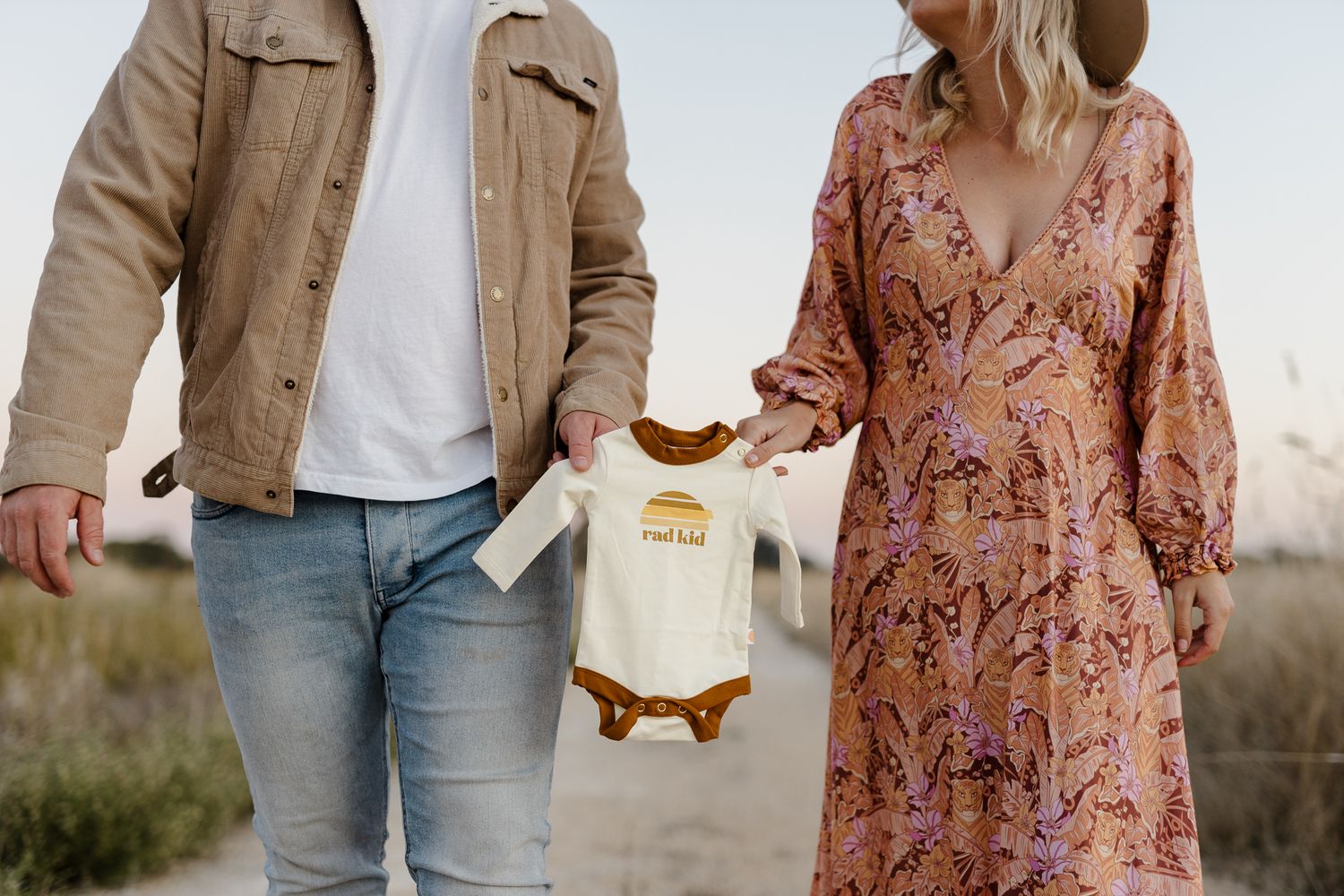 Couple displaying white baby onesie between them on outdoor path.