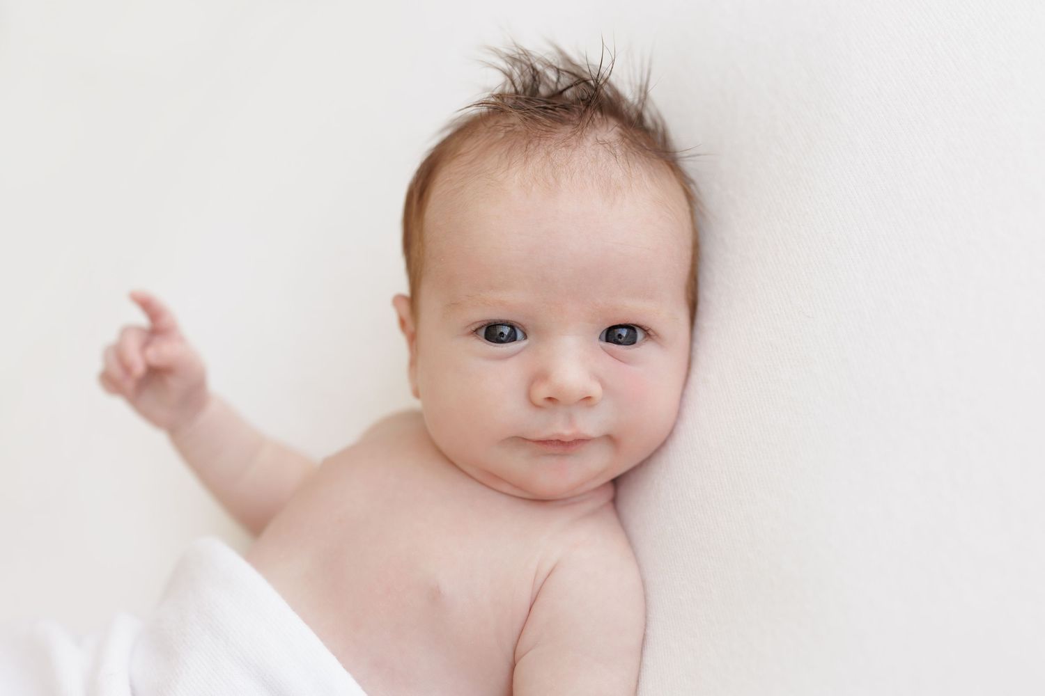 Bright-eyed baby lies on white bedding with one arm raised.