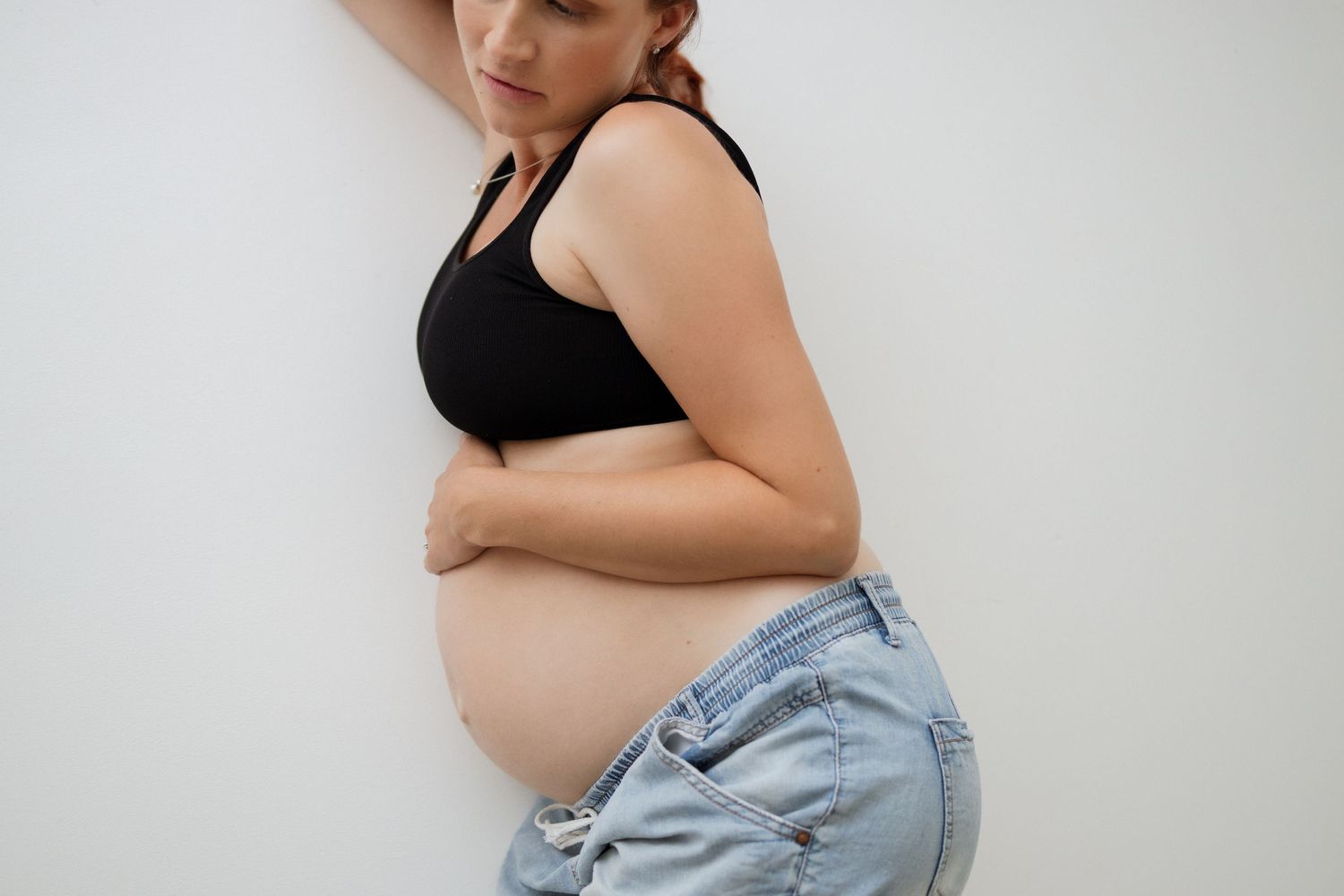 Side view of pregnant woman in black top and denim showing baby bump against white wall.
