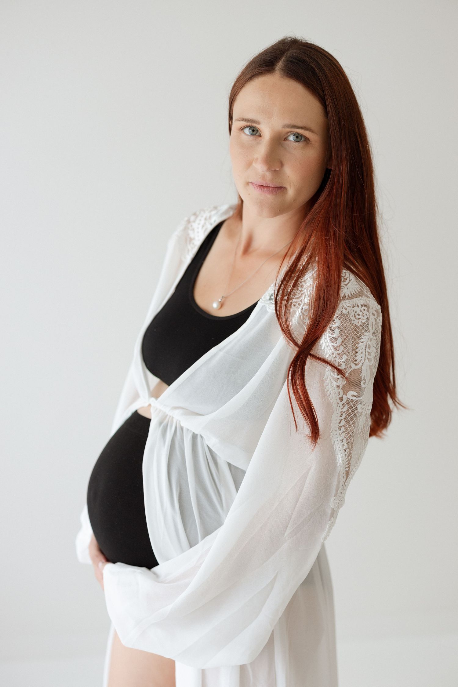 Maternity portrait featuring flowing white lace robe over black swimsuit against neutral backdrop.