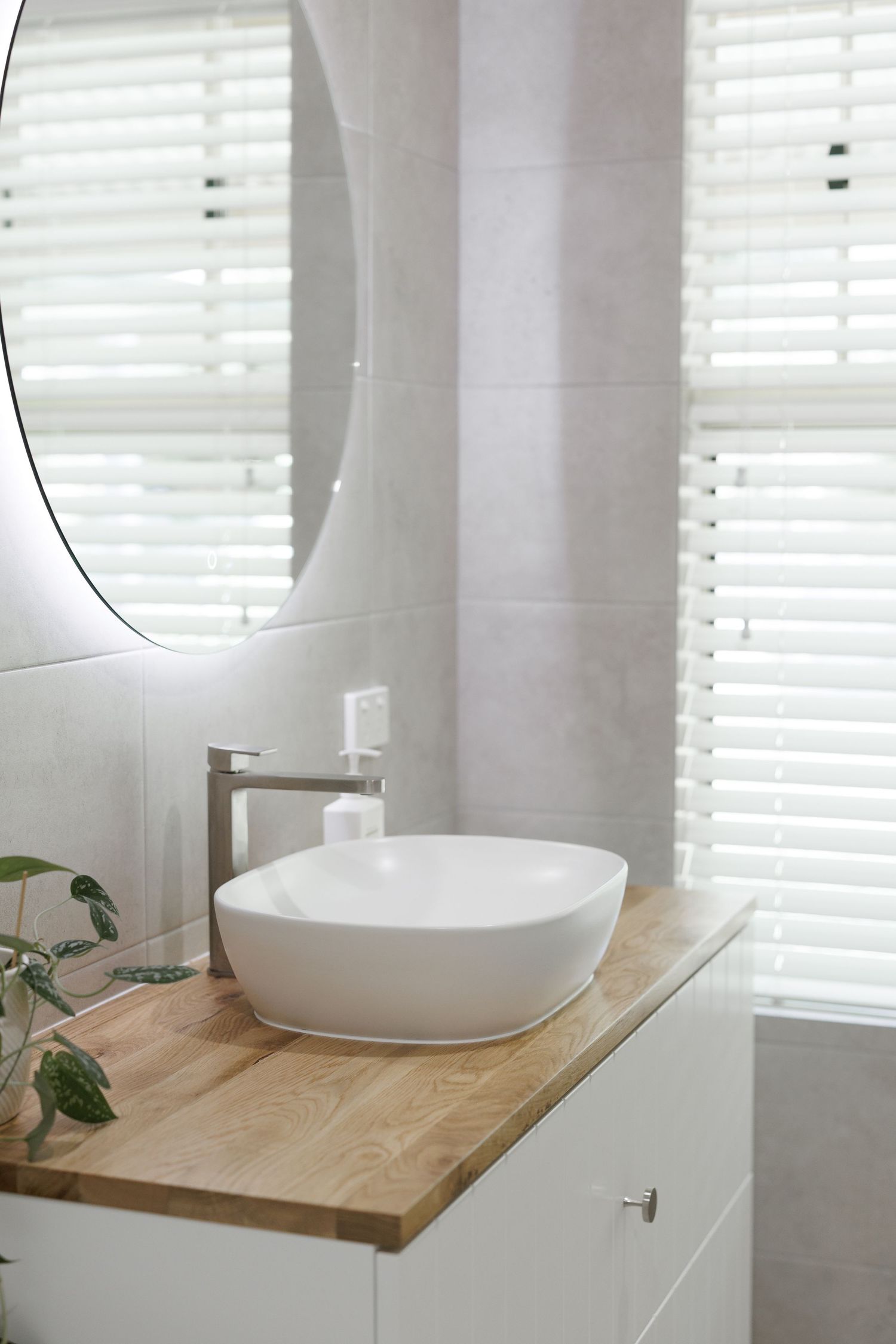 Modern white bathroom vanity with vessel sink on light wood countertop against white plantation shutters.