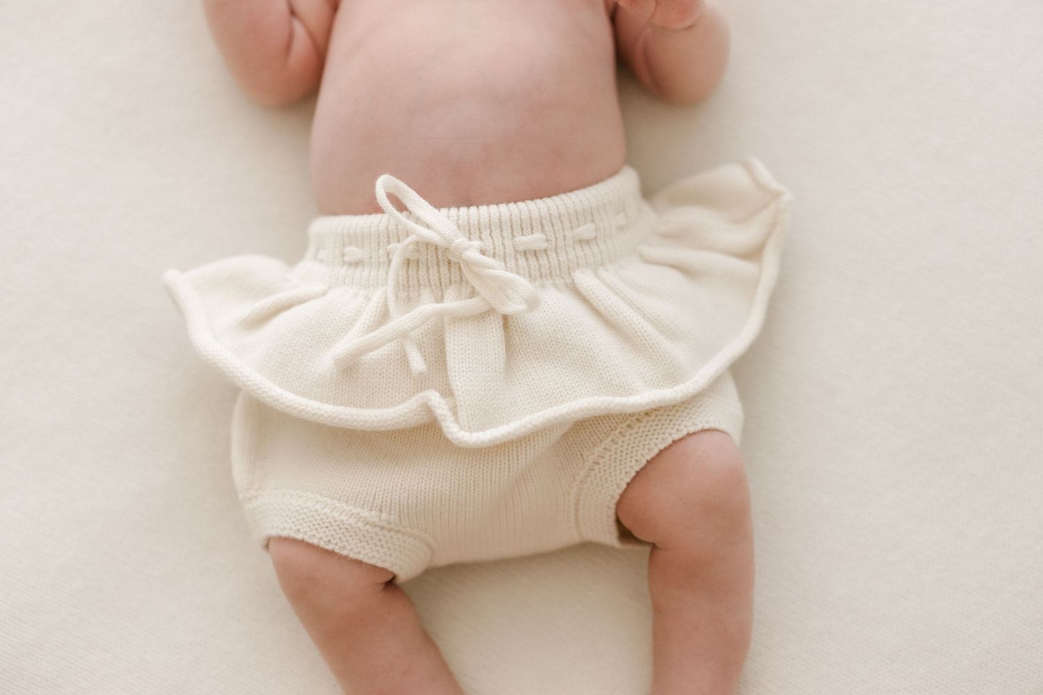 Baby wearing white ruffled bloomers lying on soft white surface.