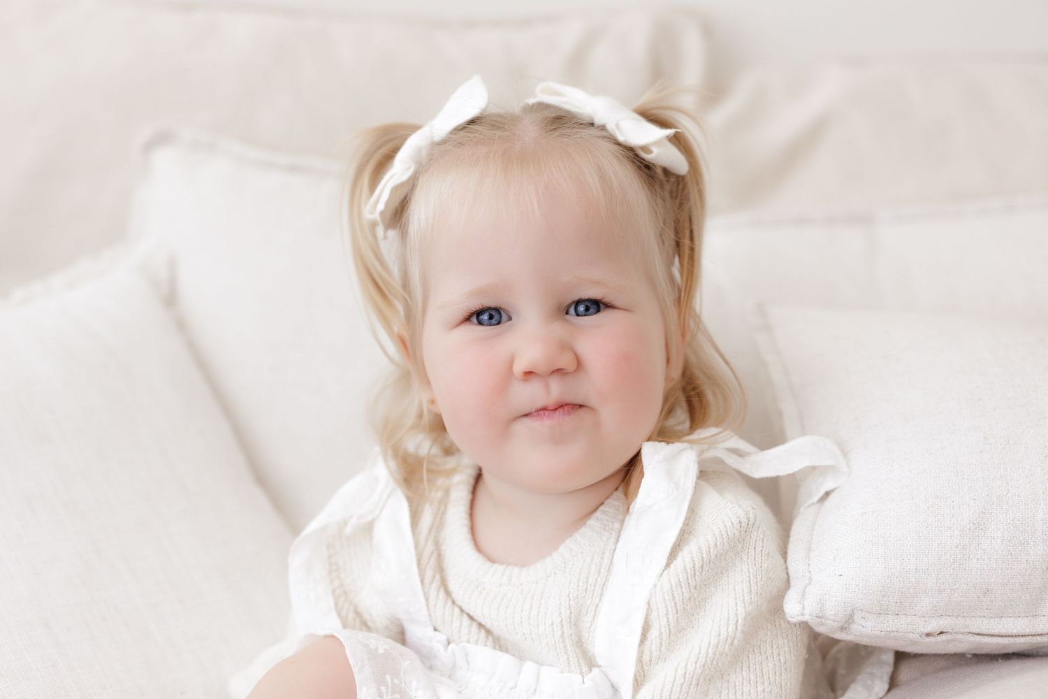 A small child in a white dress smiles while sitting on white bedding.
