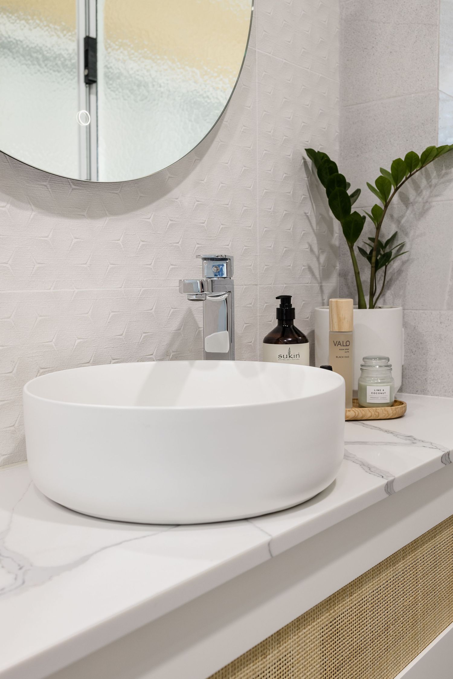 Modern white vessel sink on a wooden vanity with chrome faucet and round mirror in a minimalist bathroom design.