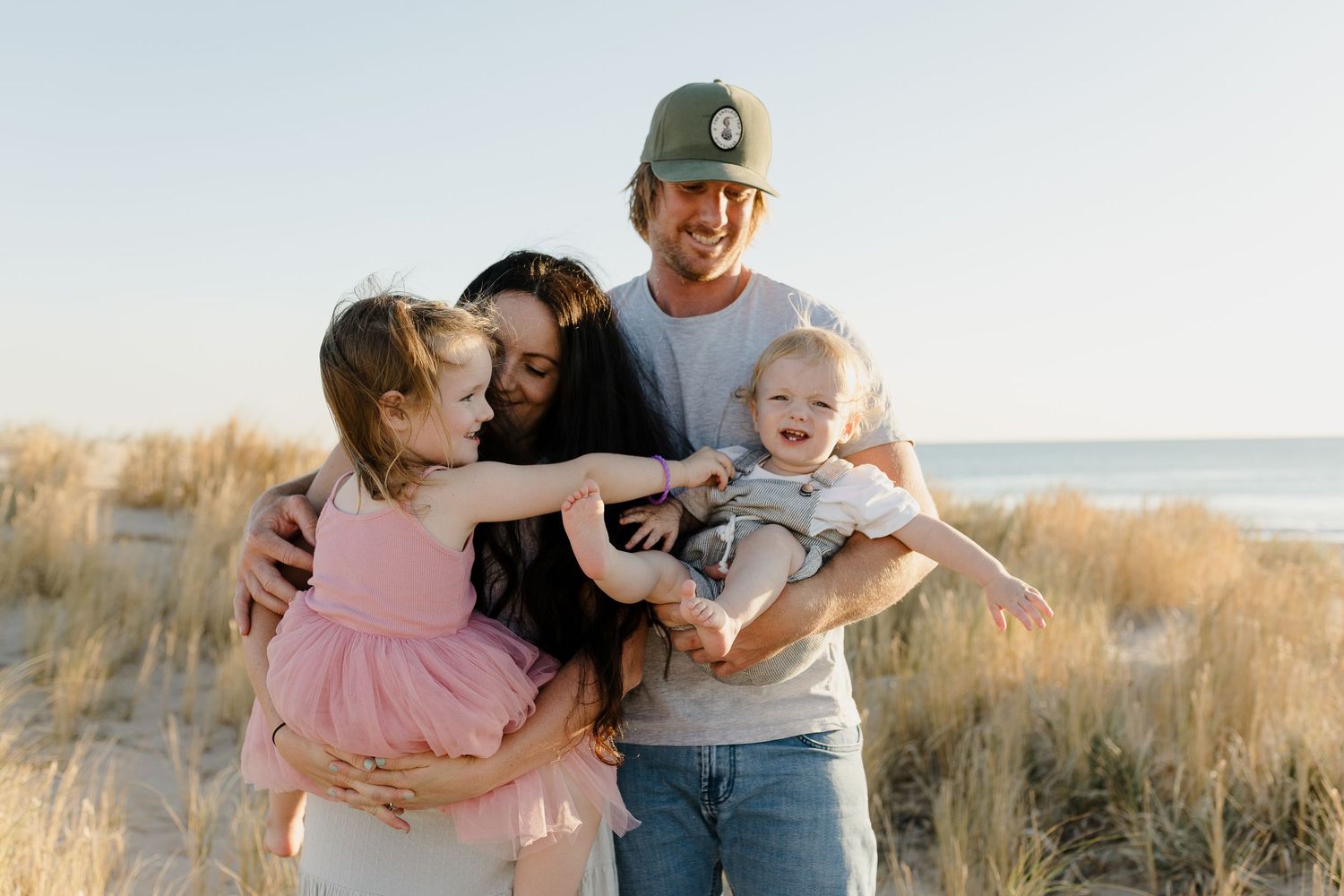 A family shares a joyful moment together on a beach with tall grass and sand dunes at sunset.