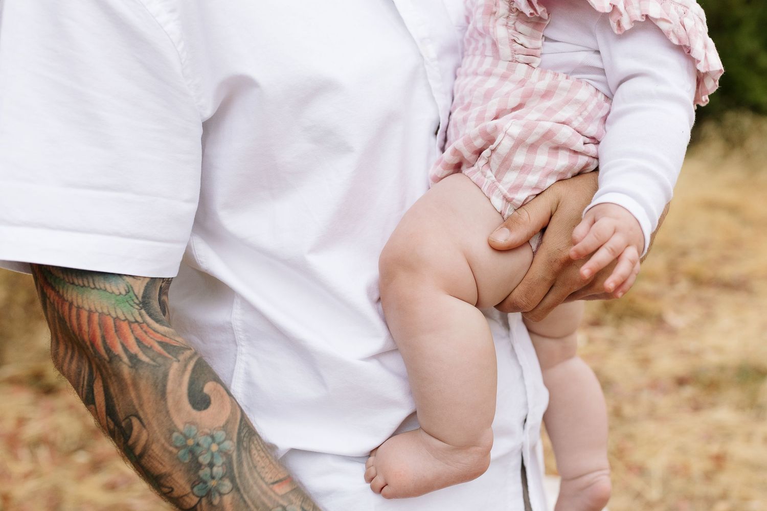 Close-up of tattooed arms holding a baby wearing pink and white clothing outdoors.