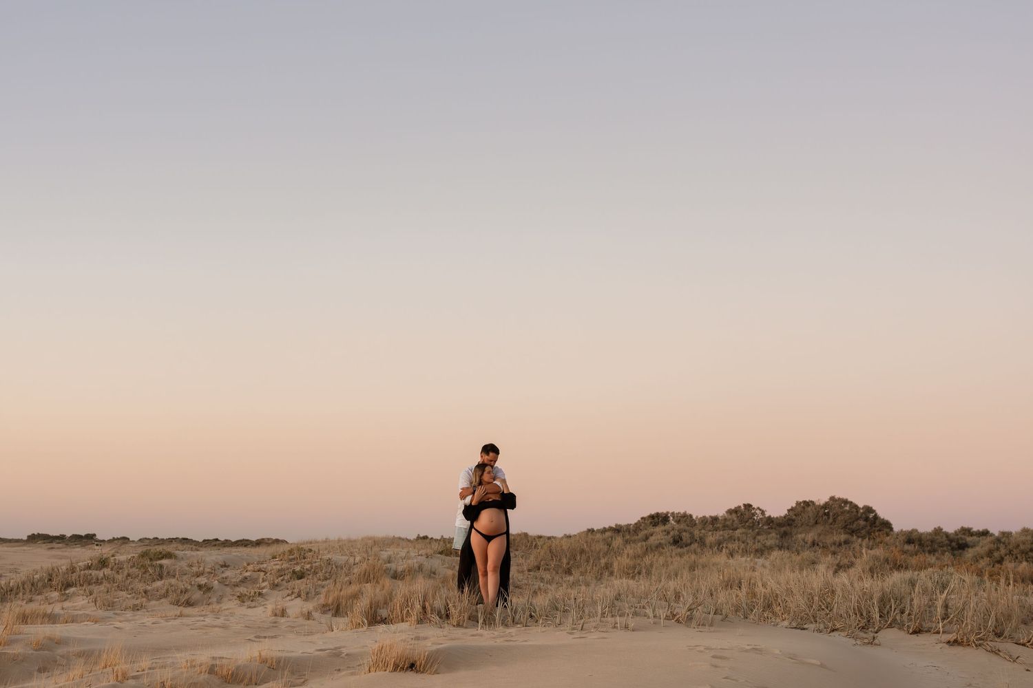 Two people embrace on coastal dunes during sunset.