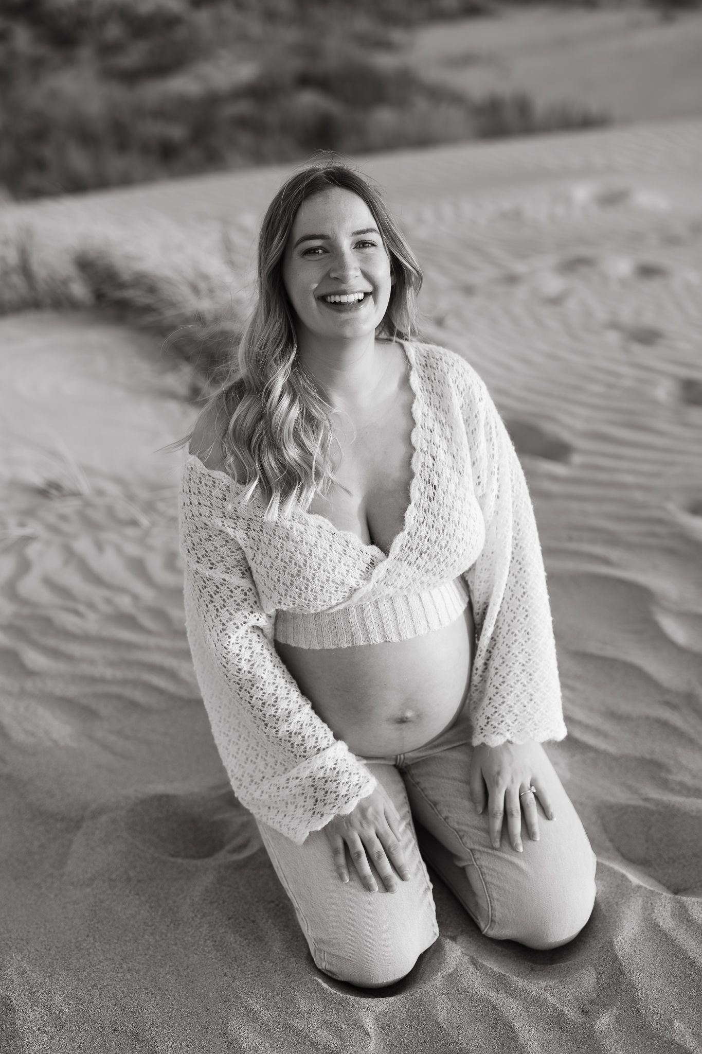 Black and white portrait of someone in a white crochet top sitting on beach sand.