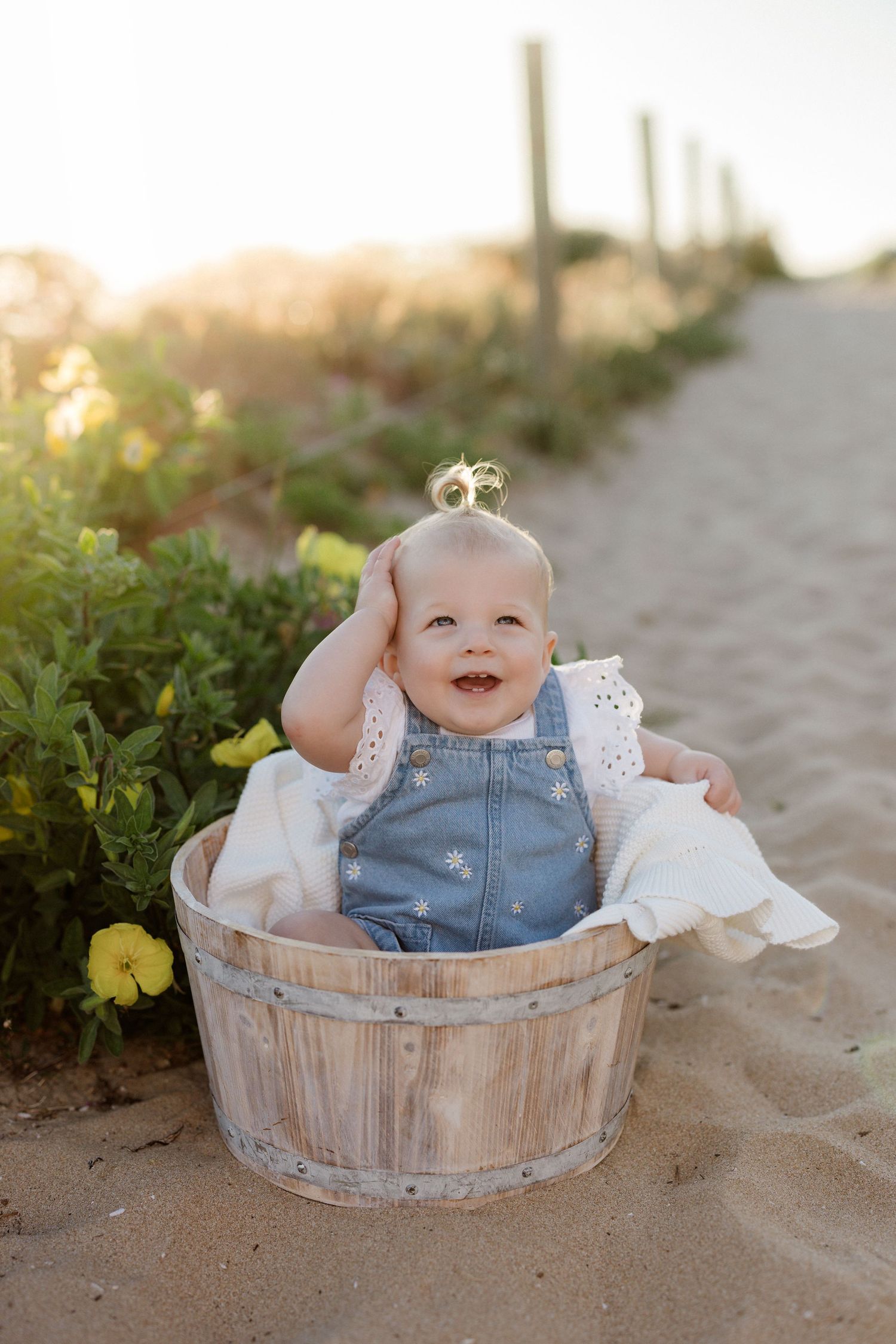 Baby in denim overalls sitting in a rustic wooden barrel on the beach at sunset.