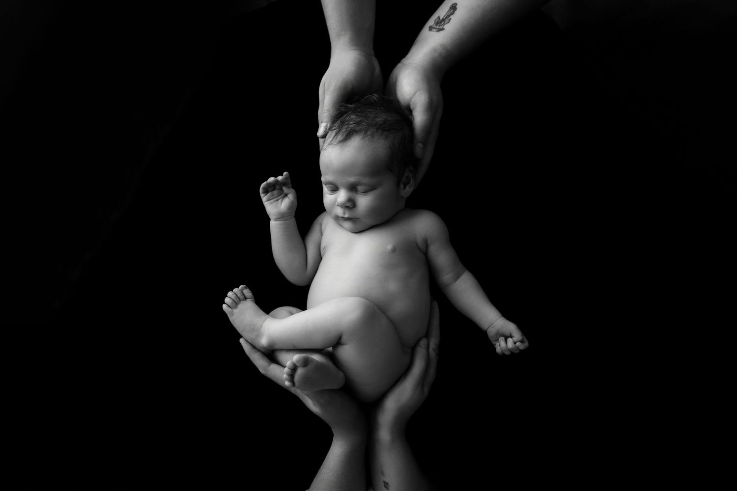 Black and white photo of a newborn baby being gently held by two pairs of adult hands against a dark background.