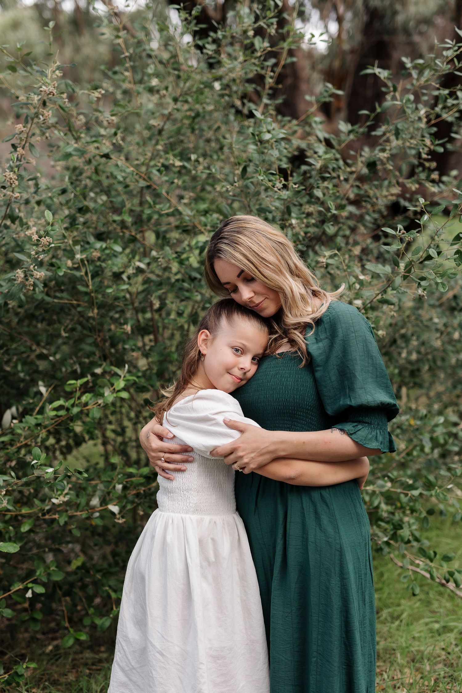 A tender outdoor embrace between two people wearing green and white dresses against a natural foliage background.