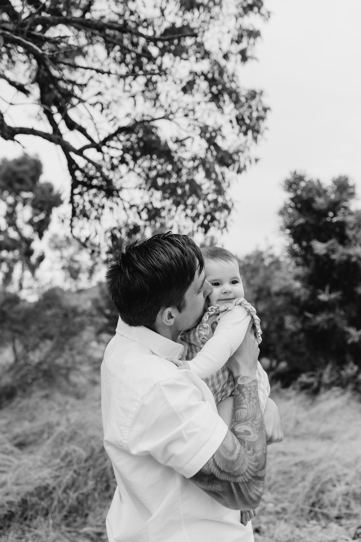 Black and white portrait of a parent holding and kissing a baby outdoors with trees in background.