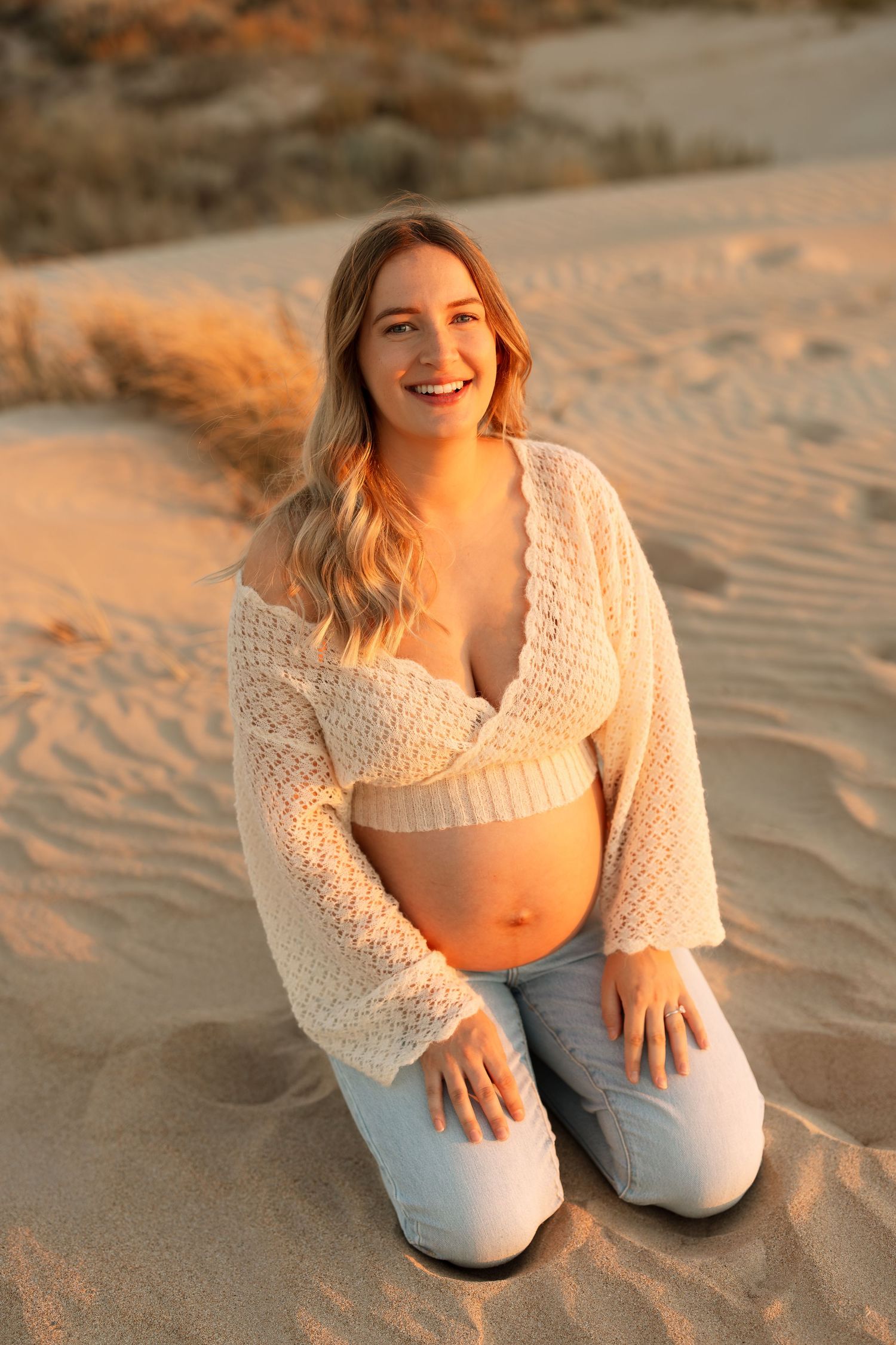 Woman wearing cream sweater and jeans poses on beach sand during golden hour.