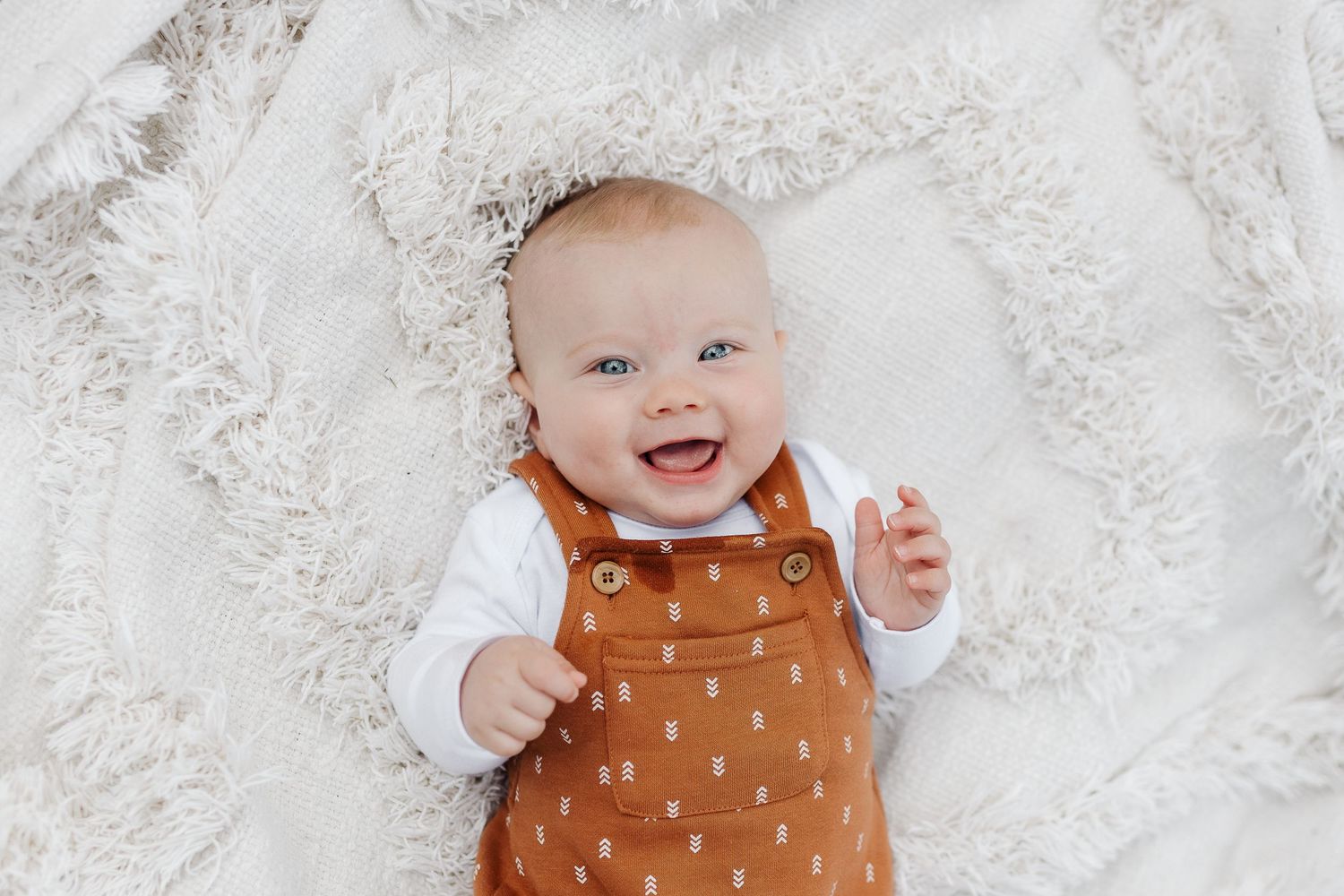 Happy baby wearing polka dot orange overalls lying on white textured blanket.