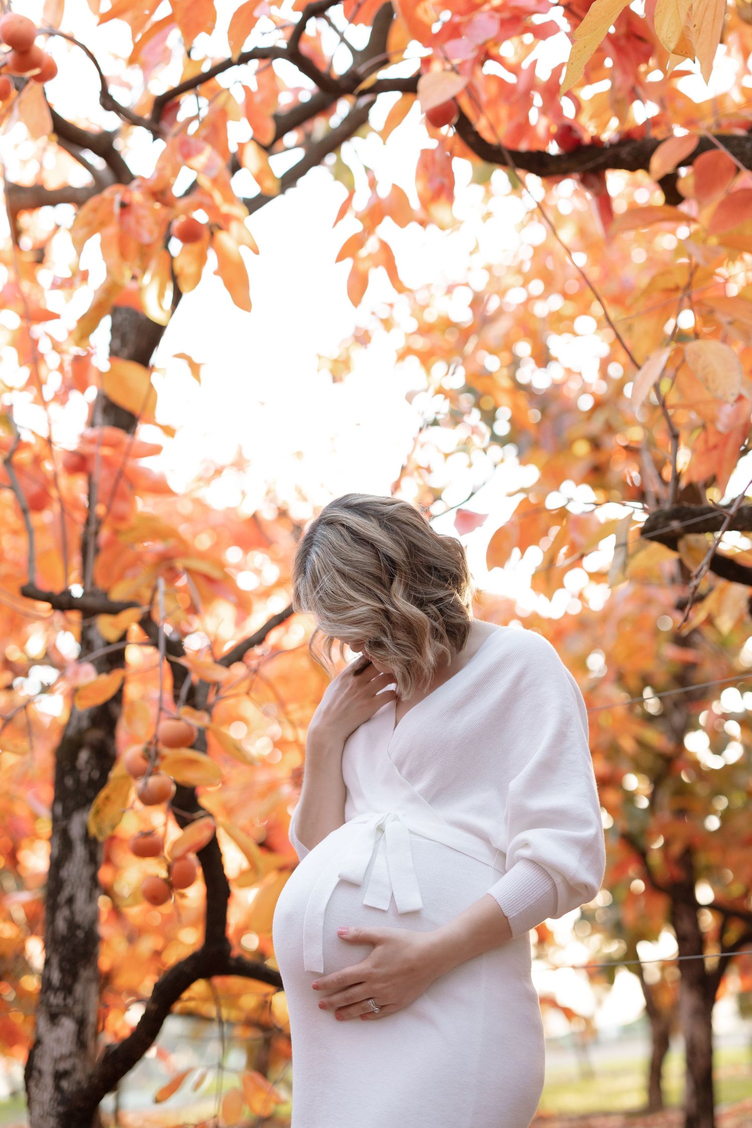 Expectant mother in white dress standing under vibrant fall trees.