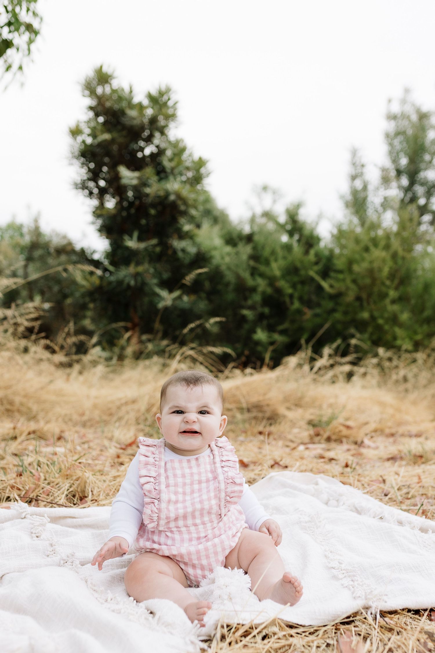Smiling baby in pink gingham romper sits on white blanket with trees in background.
