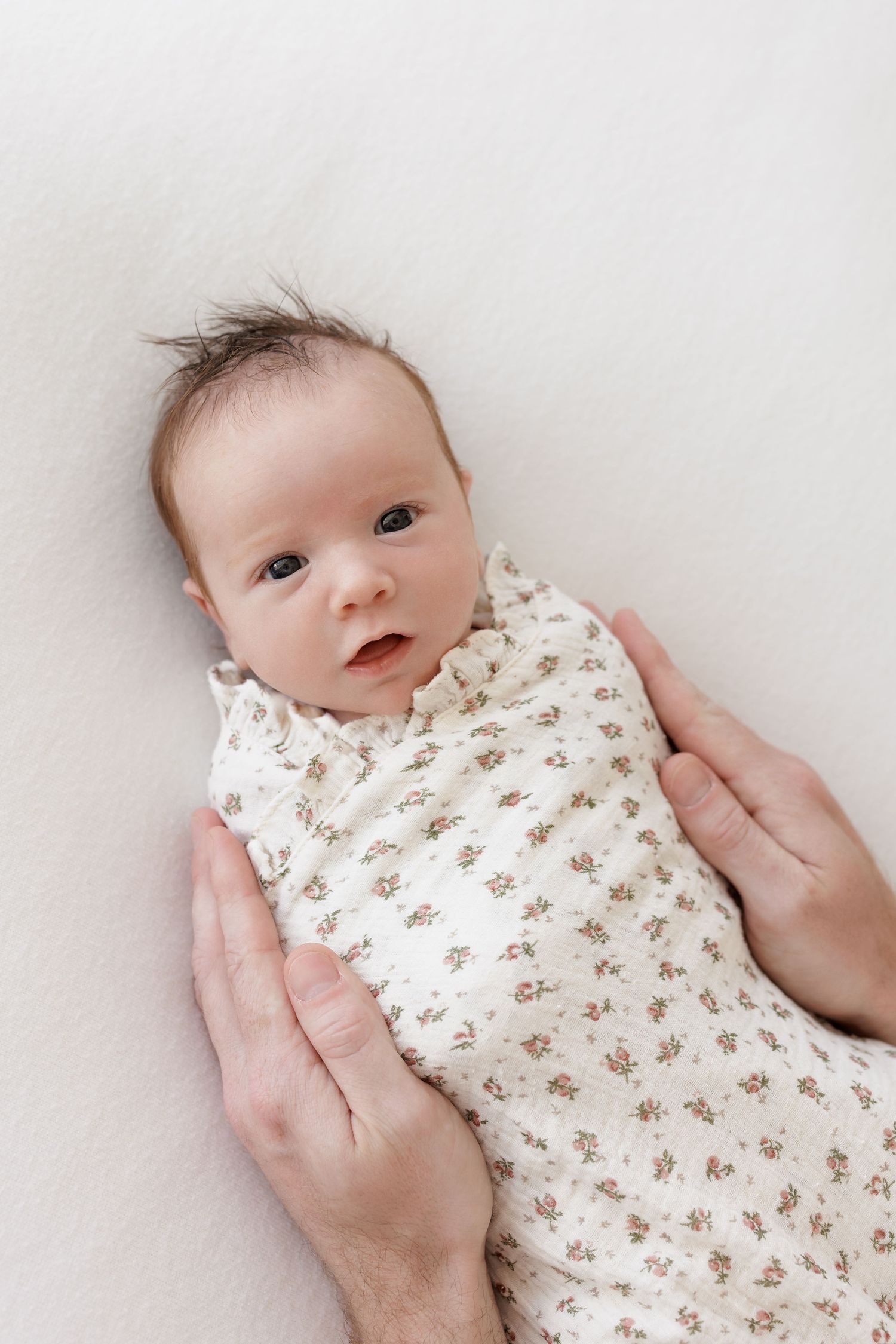 Baby in floral swaddle blanket lies on white background as hands gently support.