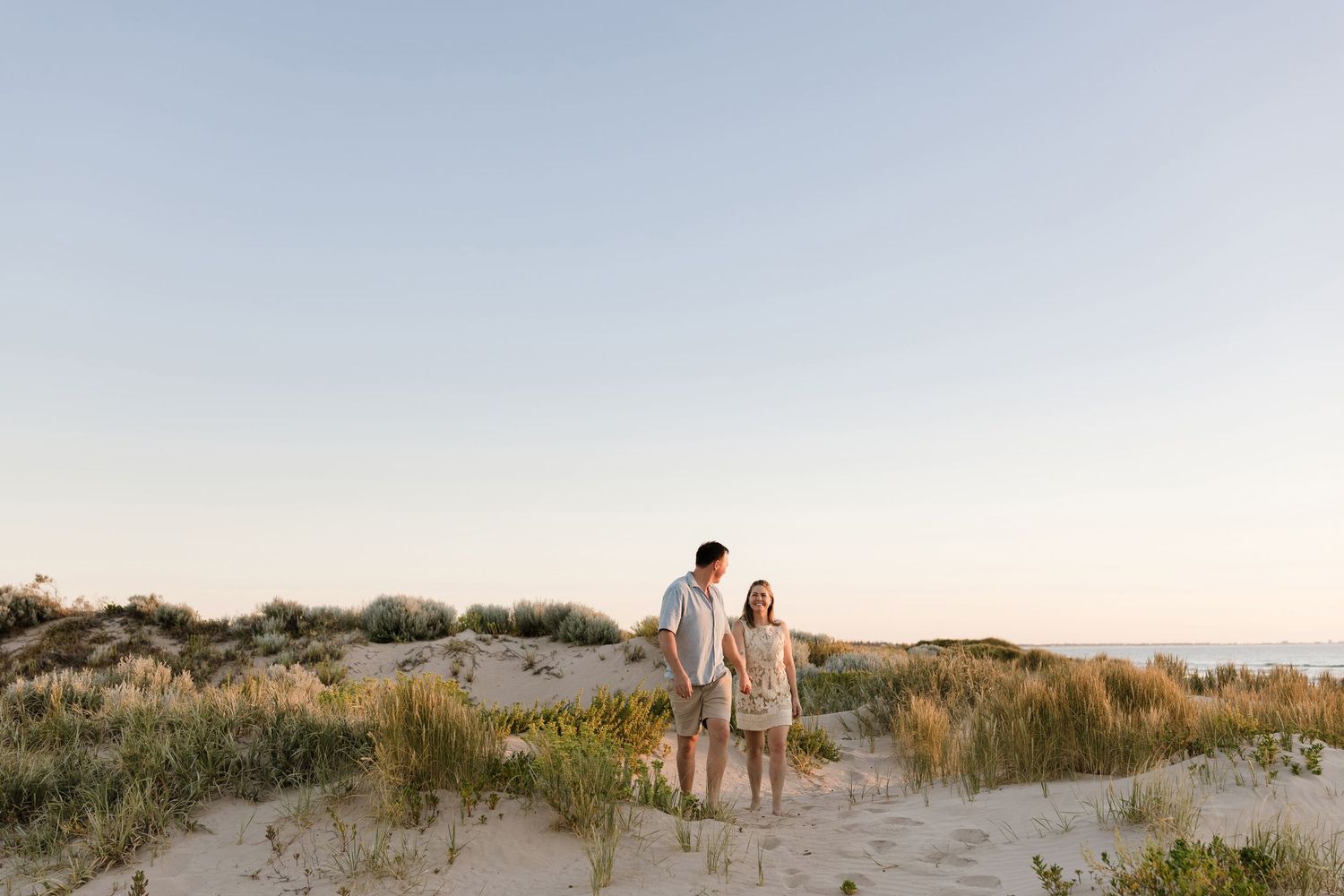 Couple holding hands on sandy beach dunes at golden hour with coastal grasses and calm ocean in the background.