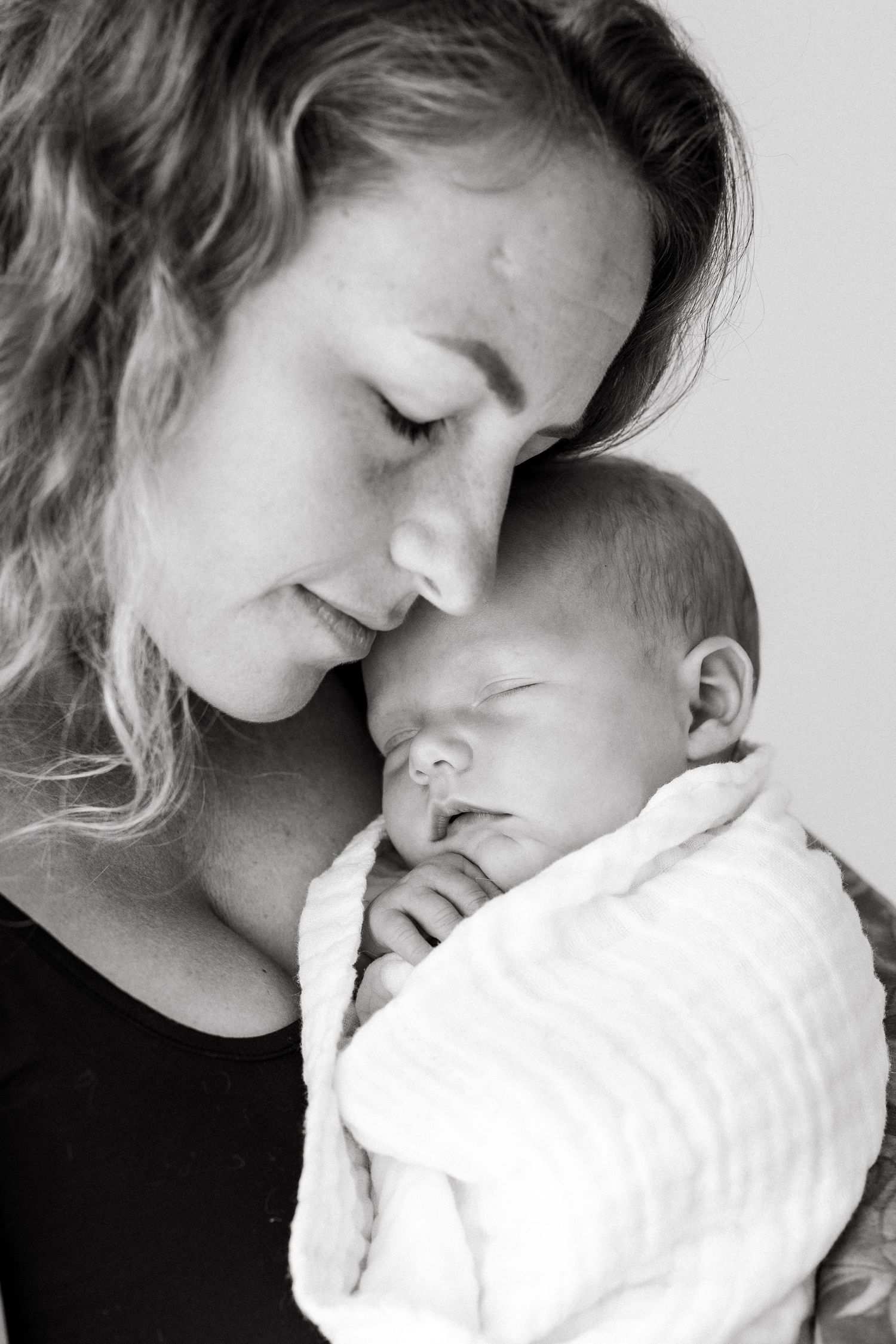 Intimate black and white portrait of a woman with wavy hair holding a sleeping newborn close.