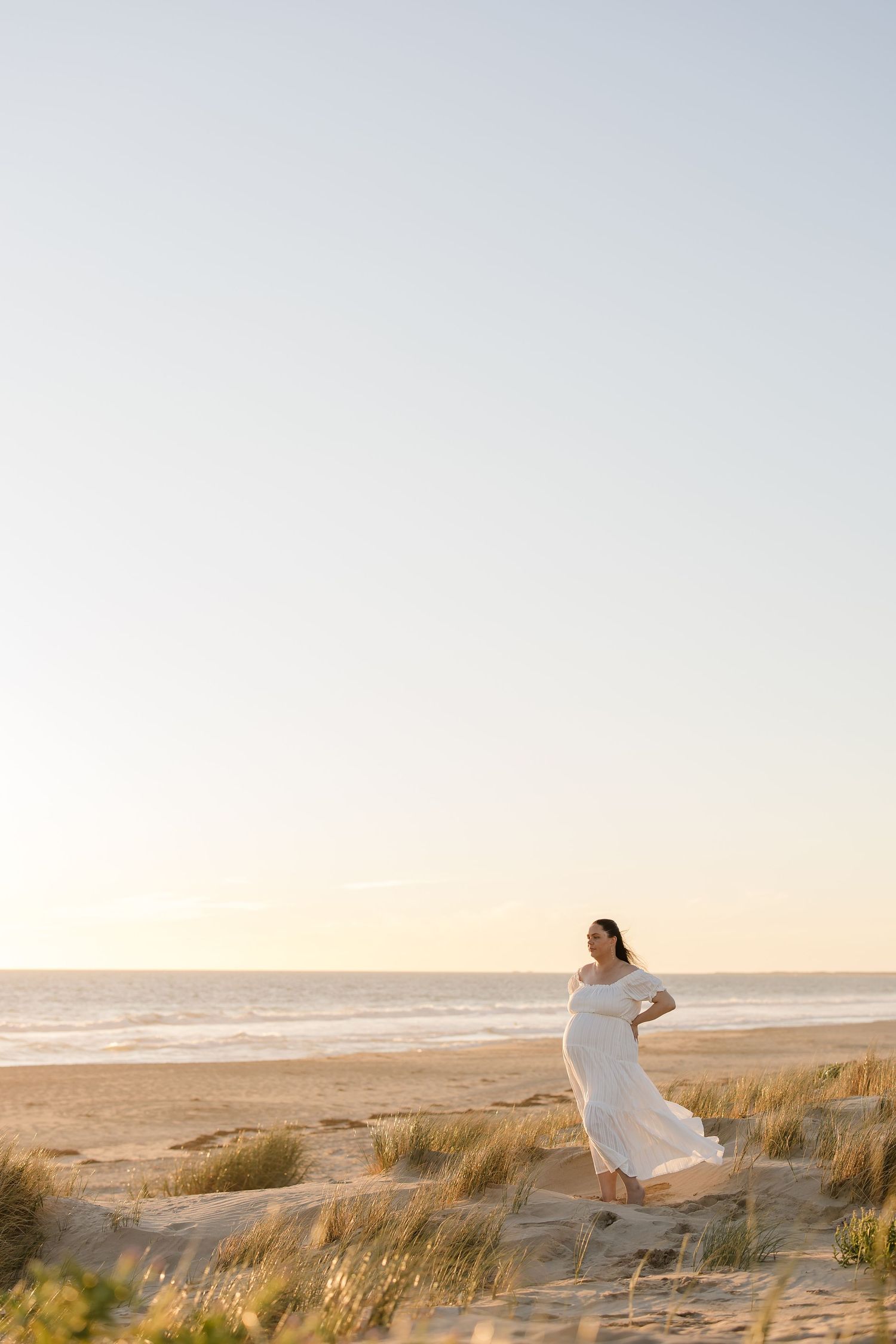Woman in flowing white dress dancing on coastal dunes at sunset.