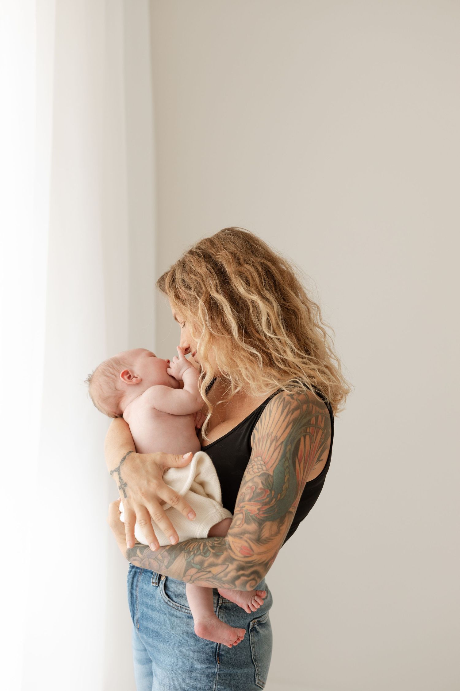 Woman with wavy hair and tattoos holding smiling baby against white wall.