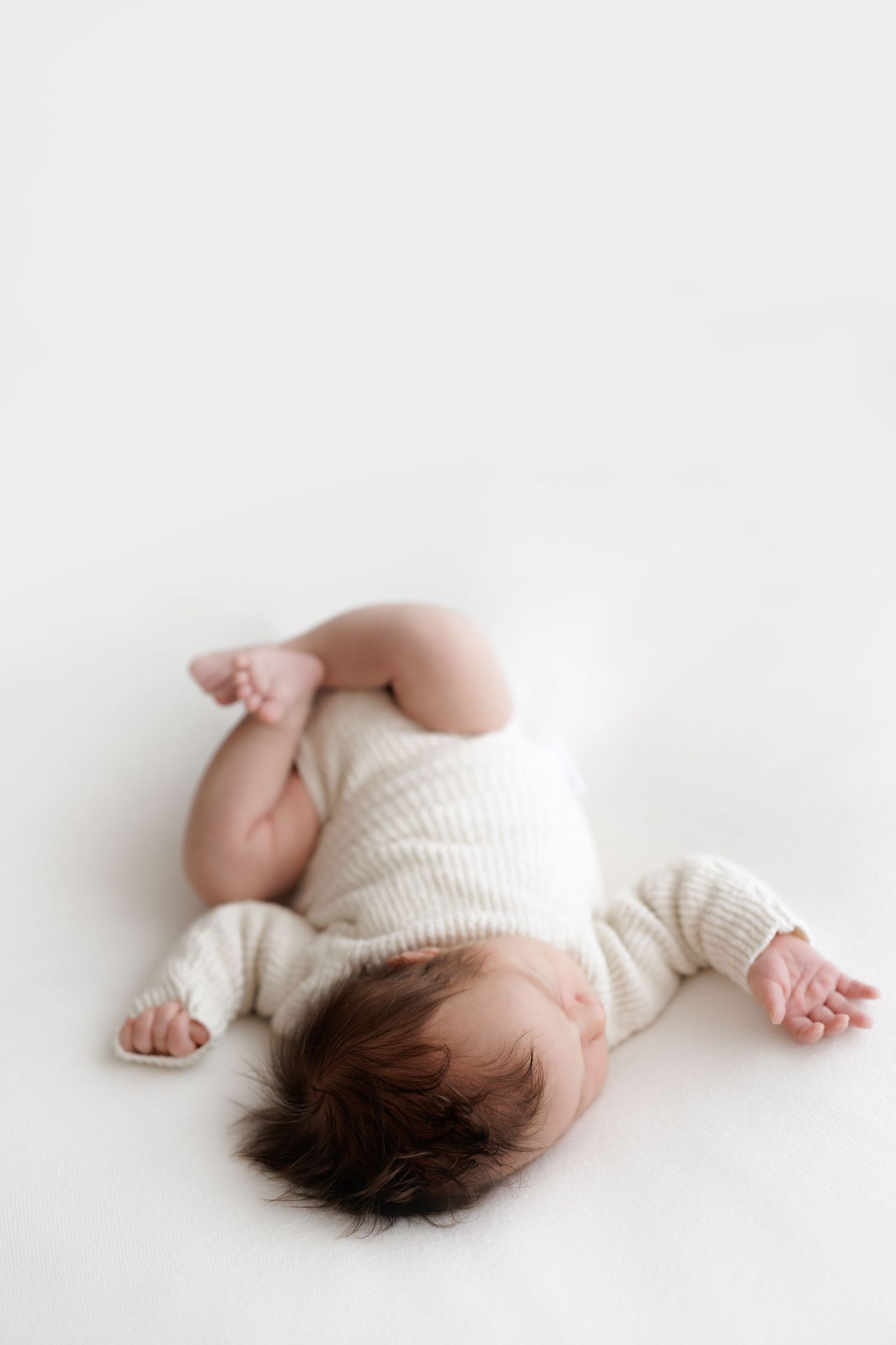 Baby in white knitted outfit lying on soft white background.