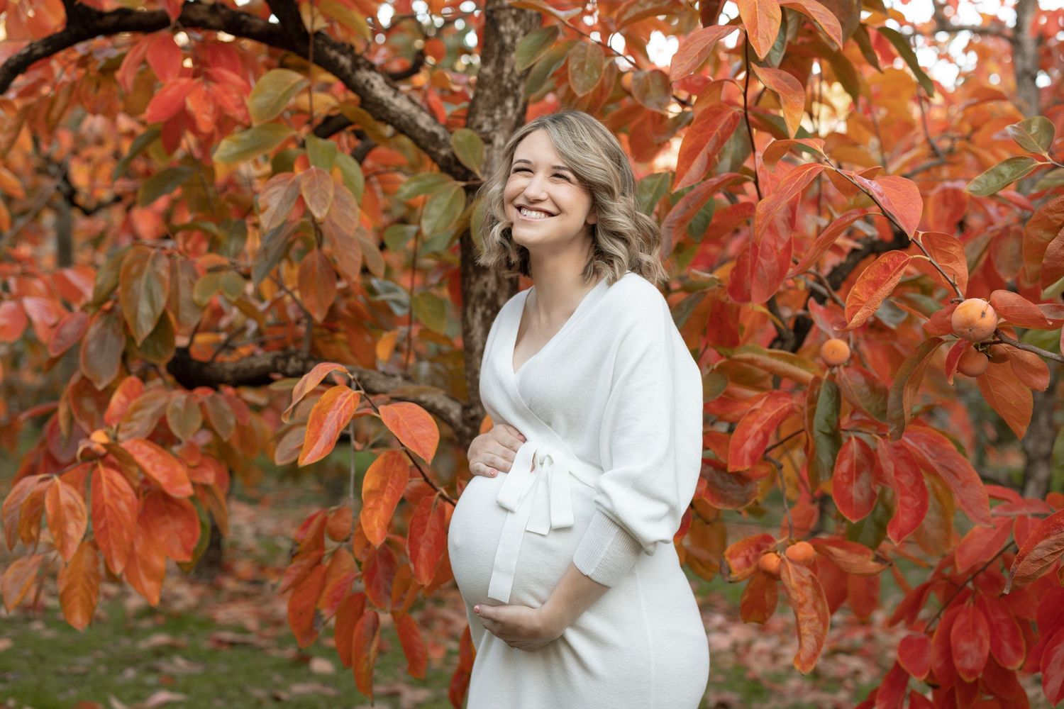 Maternity photo with white wrap dress against bright orange autumn trees.