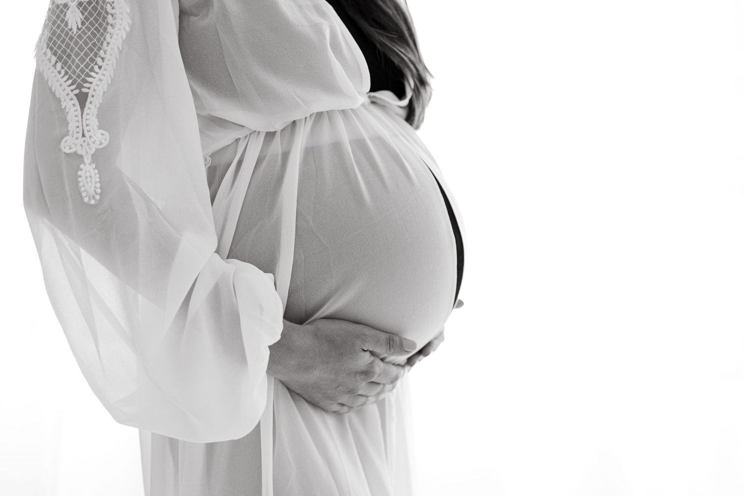 Black and white closeup of pregnant belly draped in flowing white lace maternity gown.