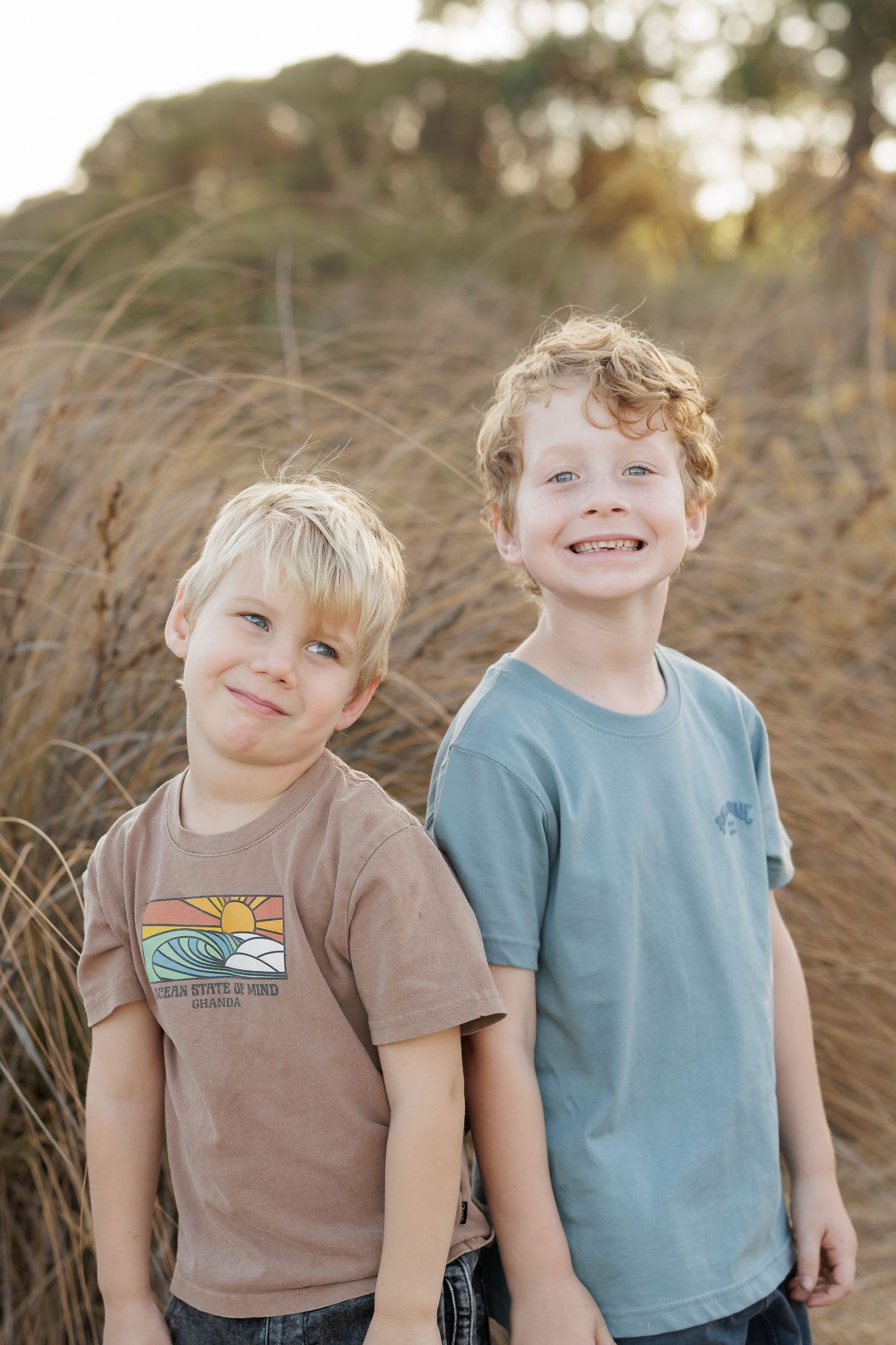 Two young boys smile outdoors with natural brush in background.