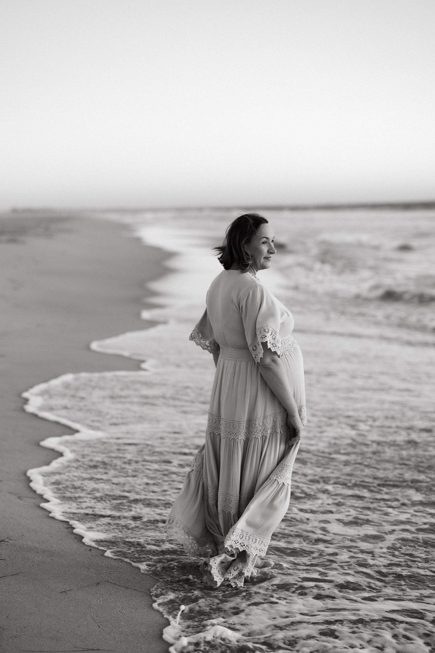 A person in a flowing white dress wades through ocean waves in black and white.