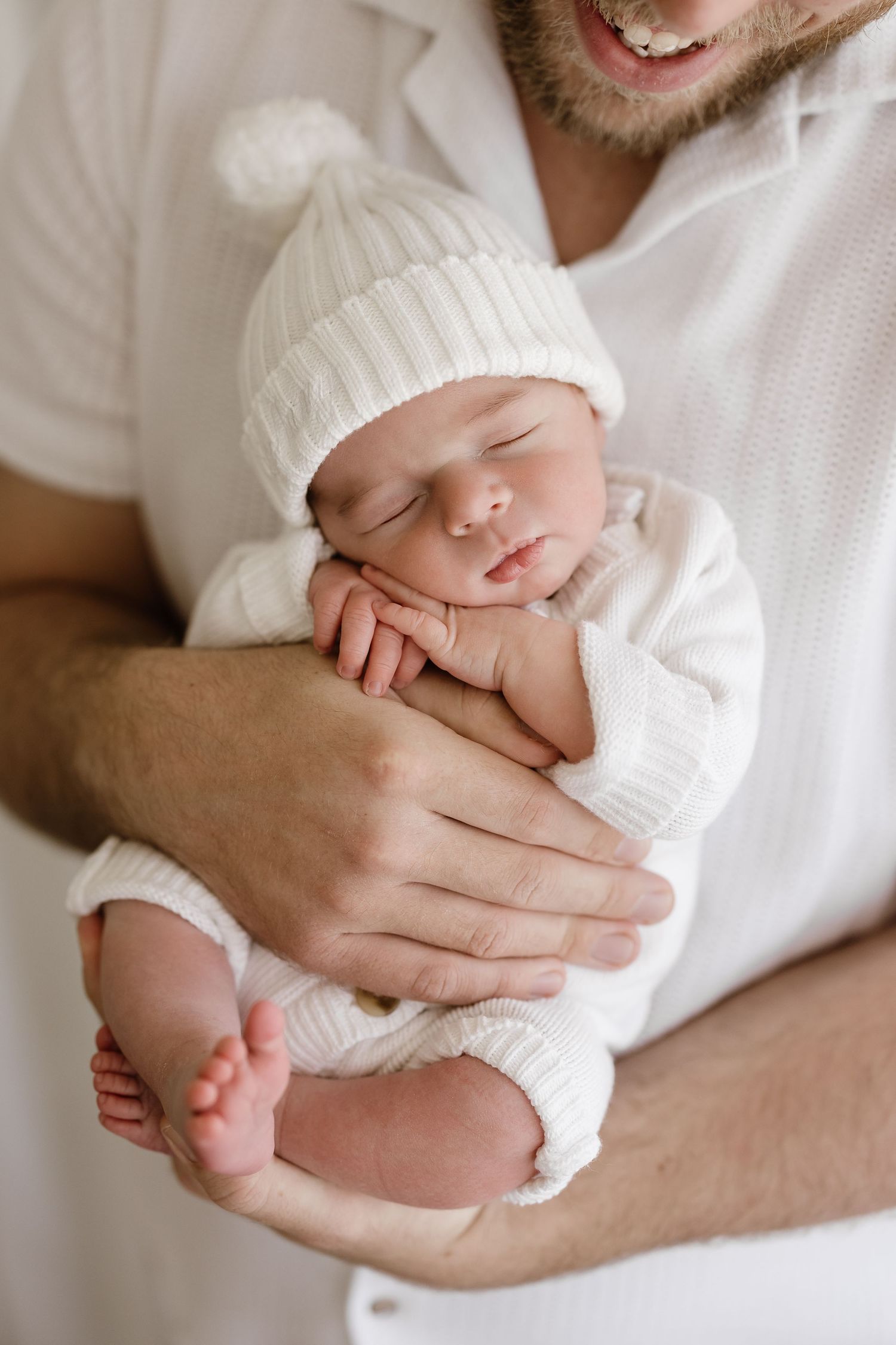 A tender moment showing hands gently cradling a sleeping newborn baby wearing a white knit hat.