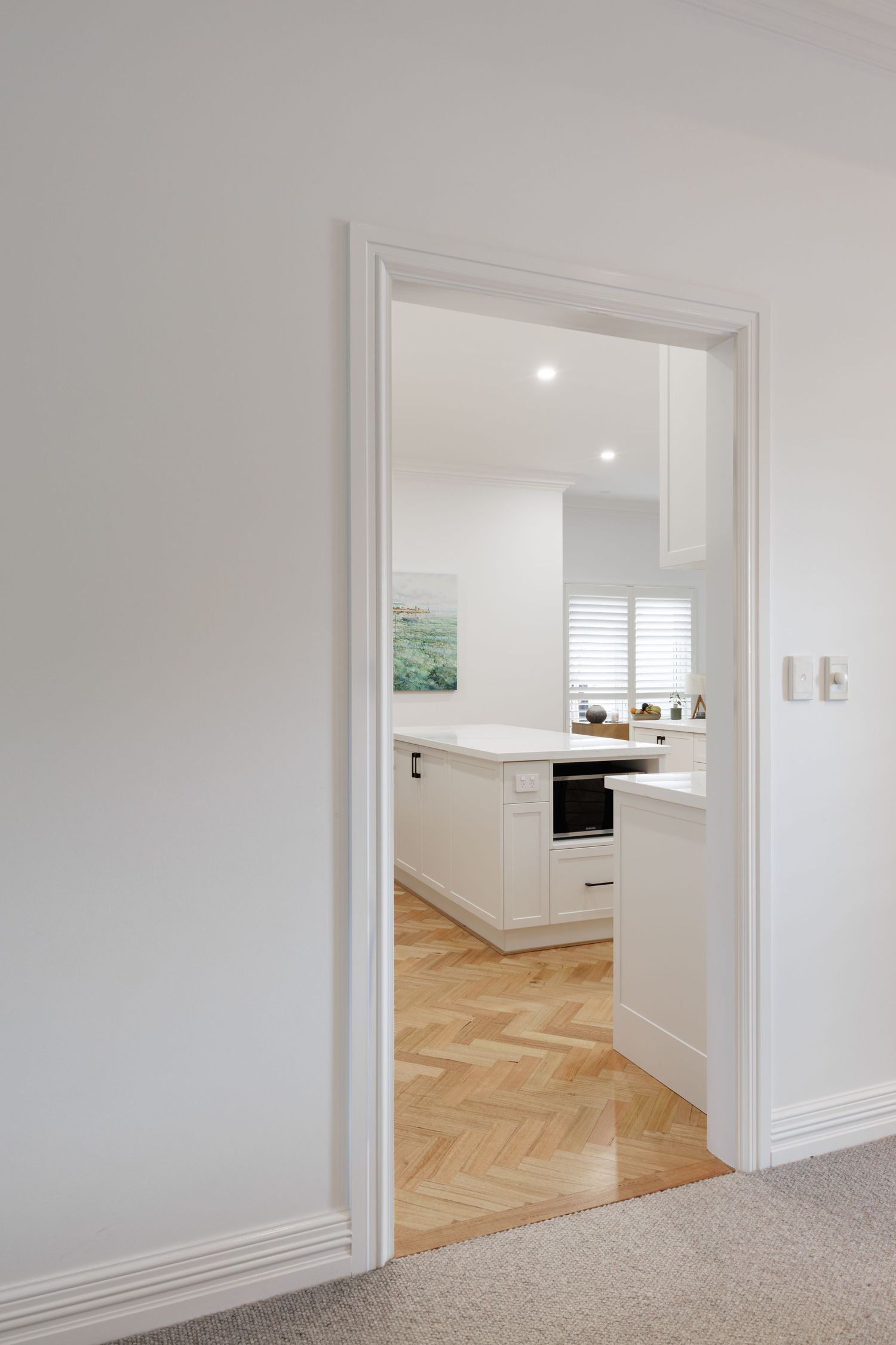 Modern white kitchen viewed through doorway with herringbone timber floors and recessed lighting.