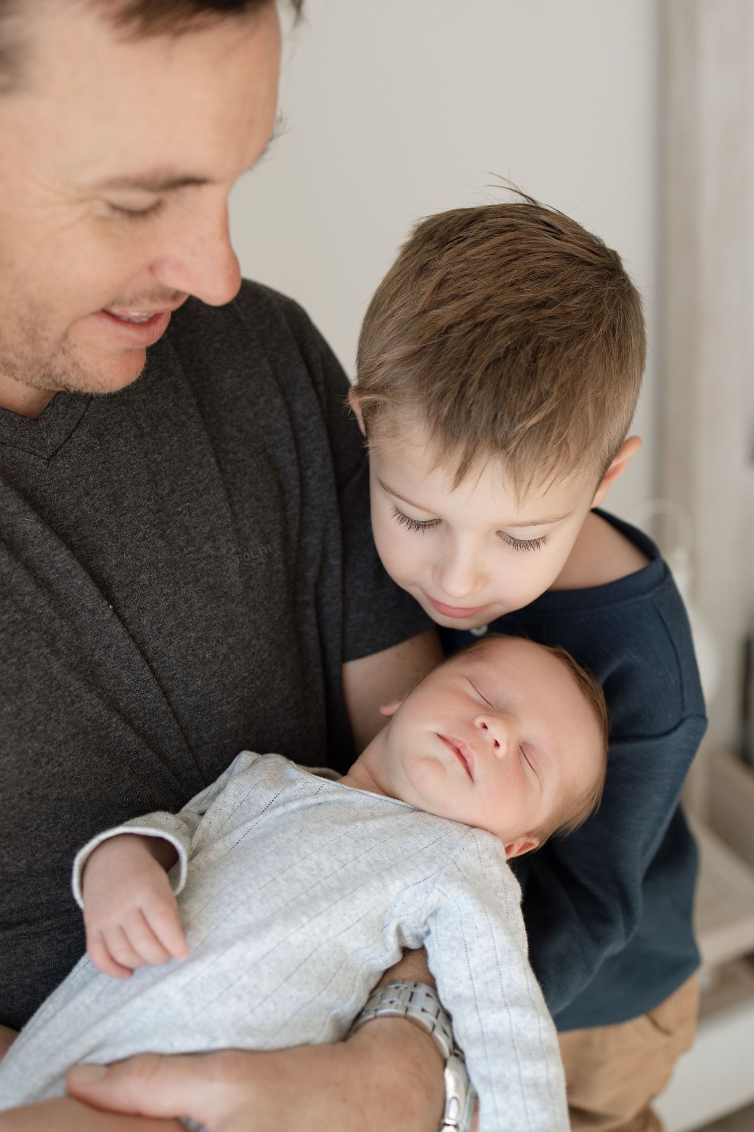 A tender family moment showing a newborn baby being held while an older sibling leans in close with care.