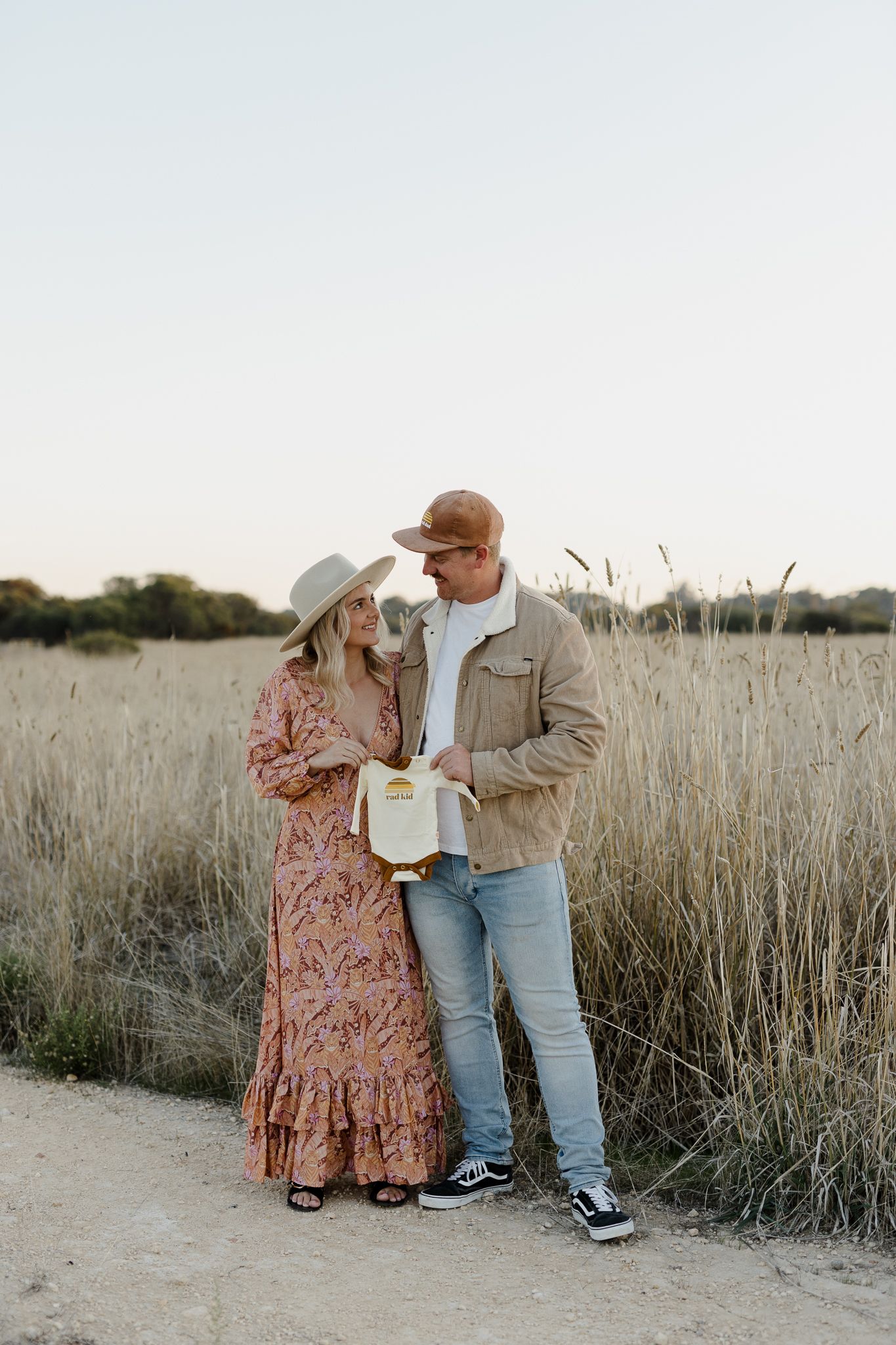 Couple sharing intimate moment while holding baby onesie on nature path.