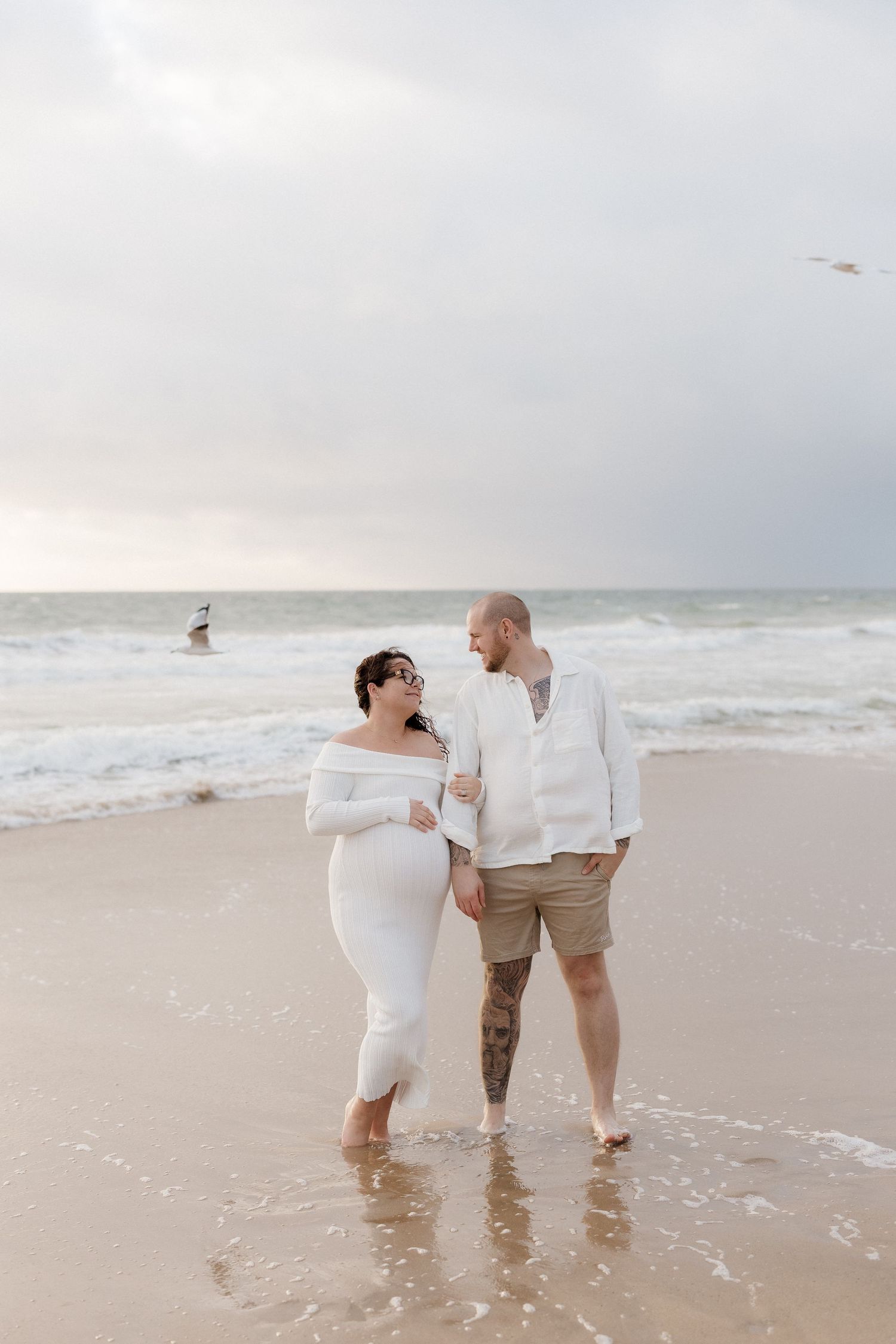 Two people in white outfits play in the ocean waves on a cloudy beach day.