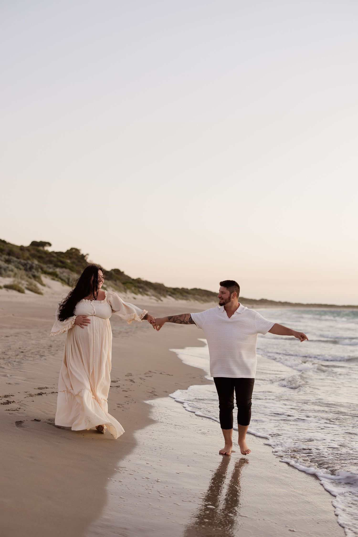 Couple in white clothing dancing and twirling together on beach at sunset.
