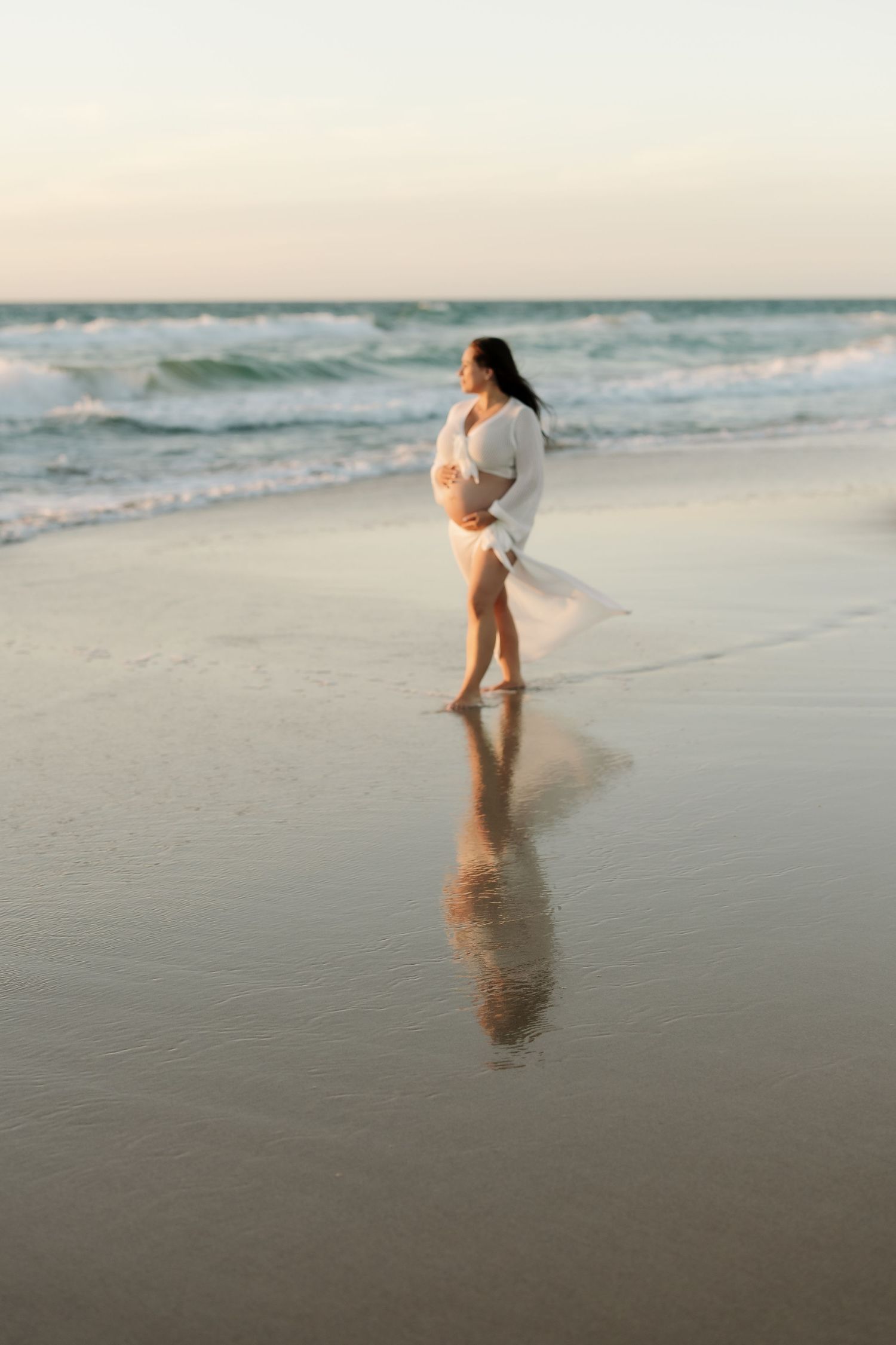 A person in a white dress walks along the shoreline at sunset as waves gently roll onto the sandy beach.