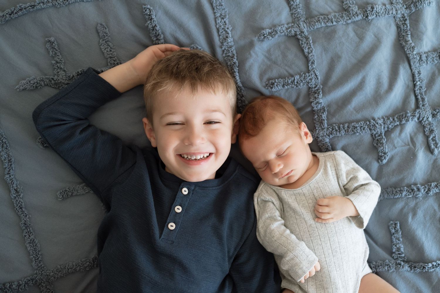 Two children lying together on a gray textured bedspread, one wearing navy and one in white.