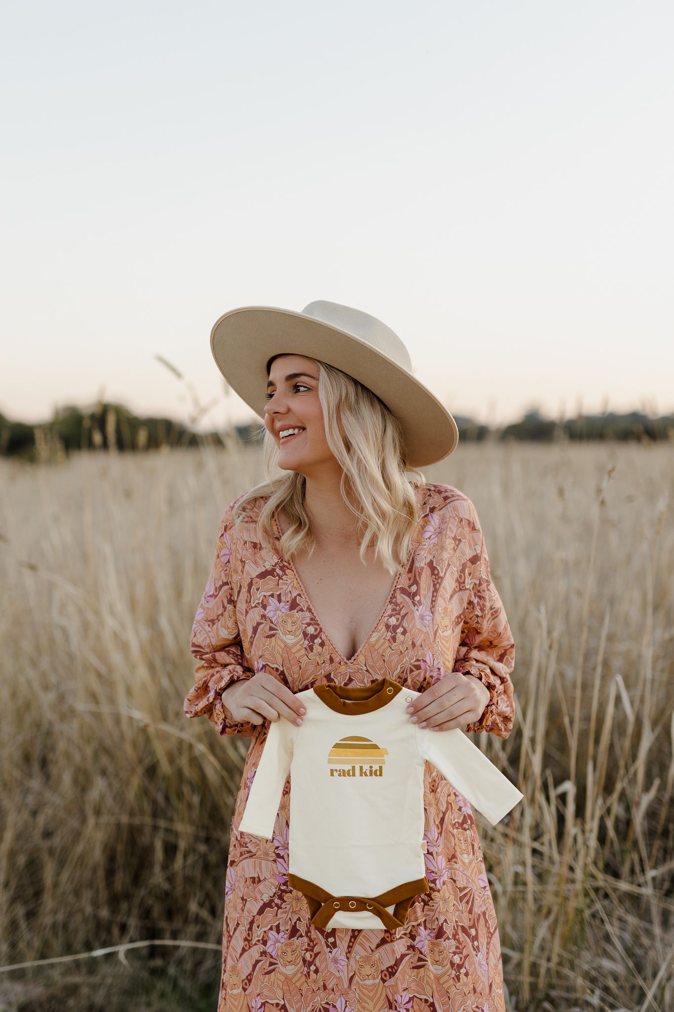 Woman in floral dress and wide brim hat holding baby onesie in field at sunset.