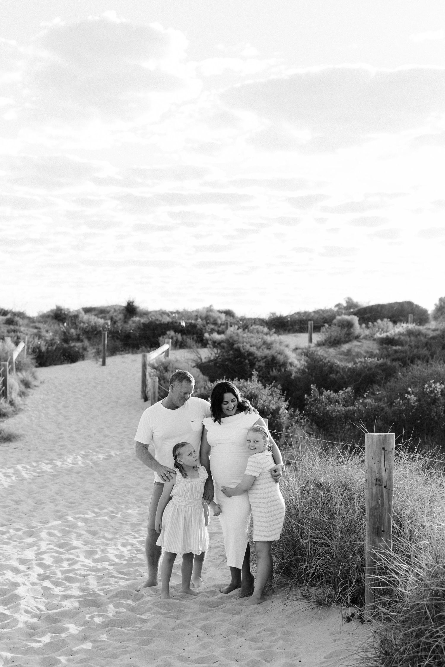 Black and white family photo on sandy beach path, parents with two young children surrounded by dune grass.