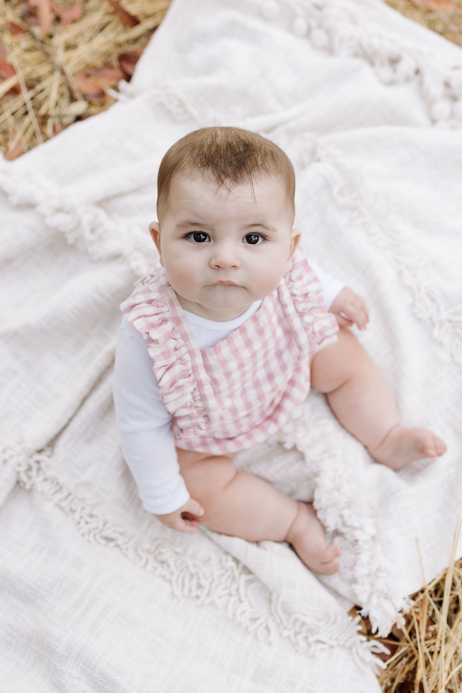 Baby wearing pink and white gingham outfit sits on lace blanket in outdoor setting.