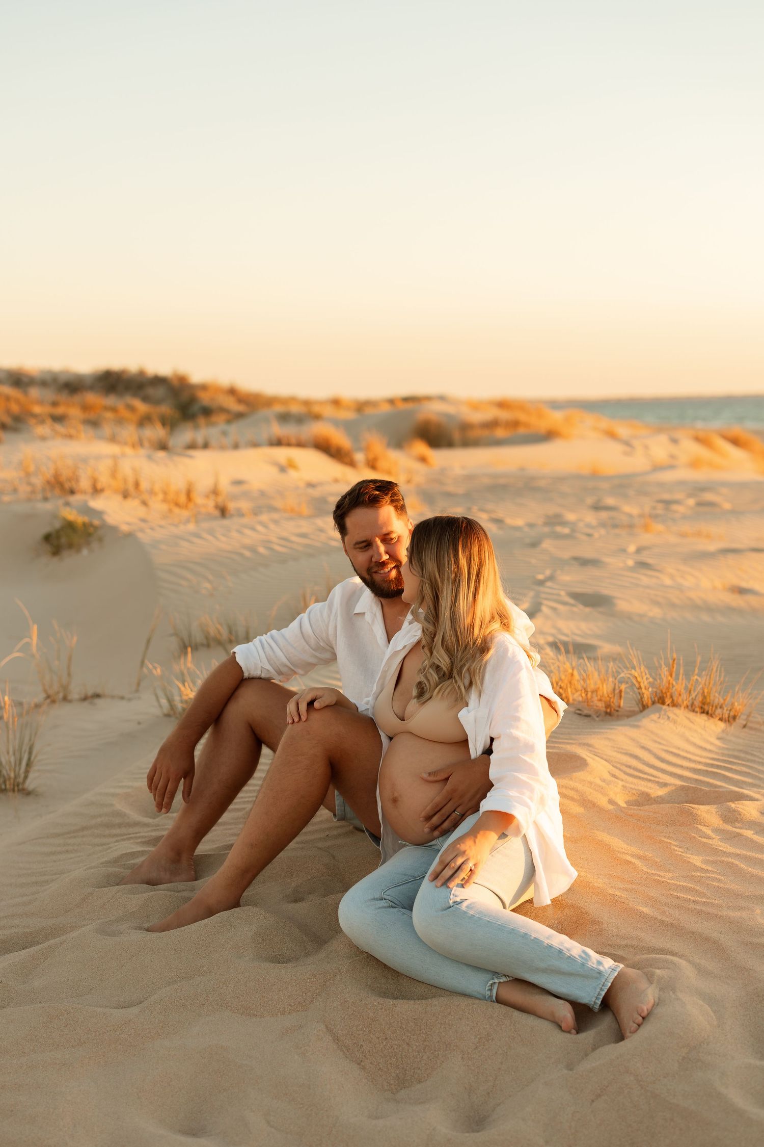 Couple sitting together on sandy beach dunes at sunset in casual white outfits.