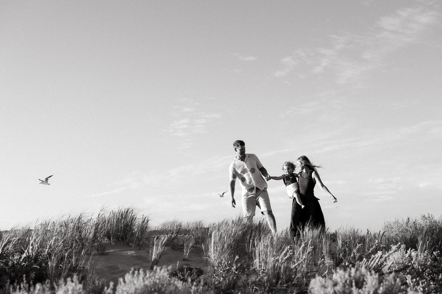 Black and white photo of two people running through coastal dunes with a bird flying in the background.